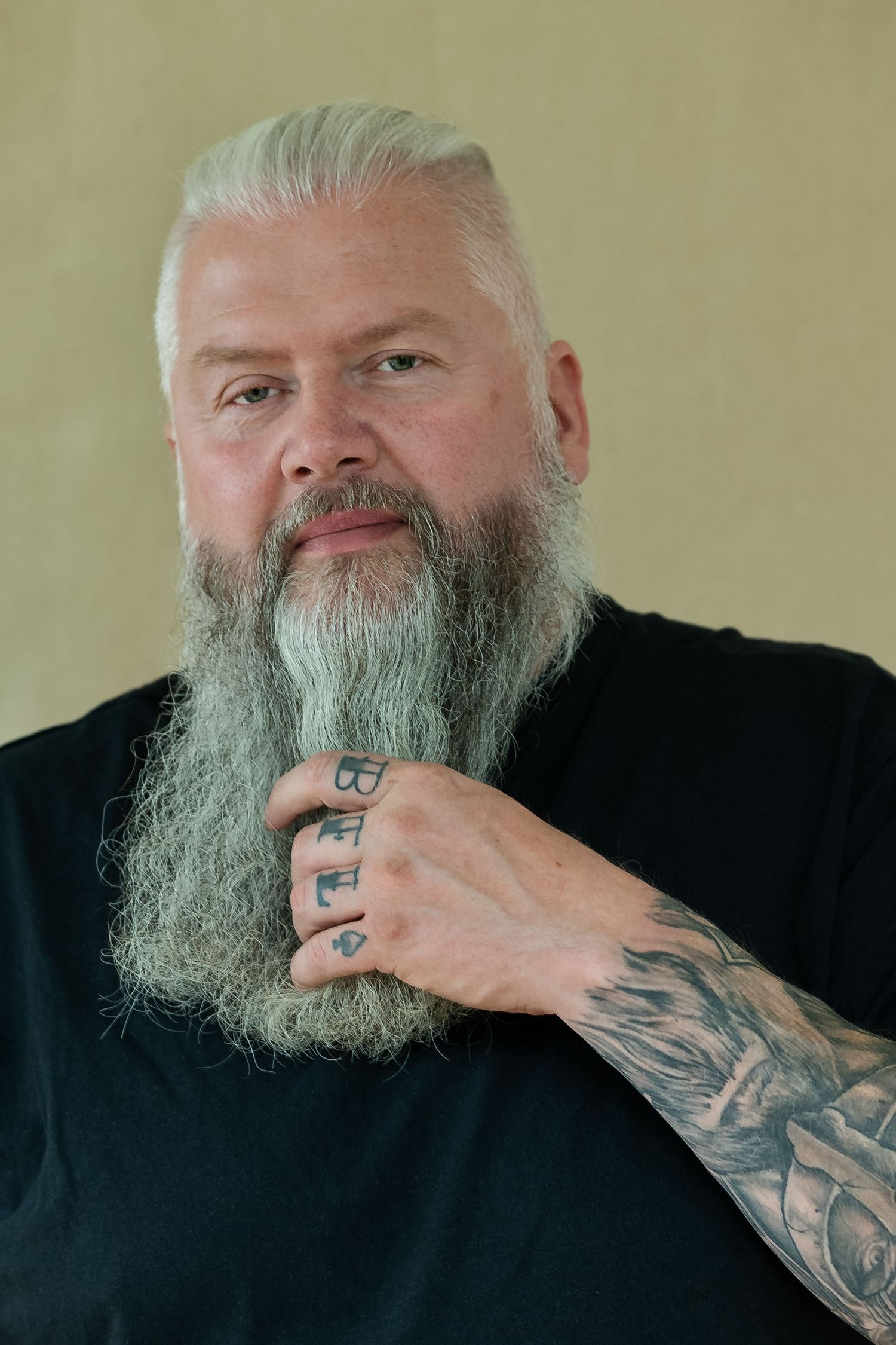 Tattooed middle-aged man with white hair and long gray beard wearing black shirt, posing with hand on chin.