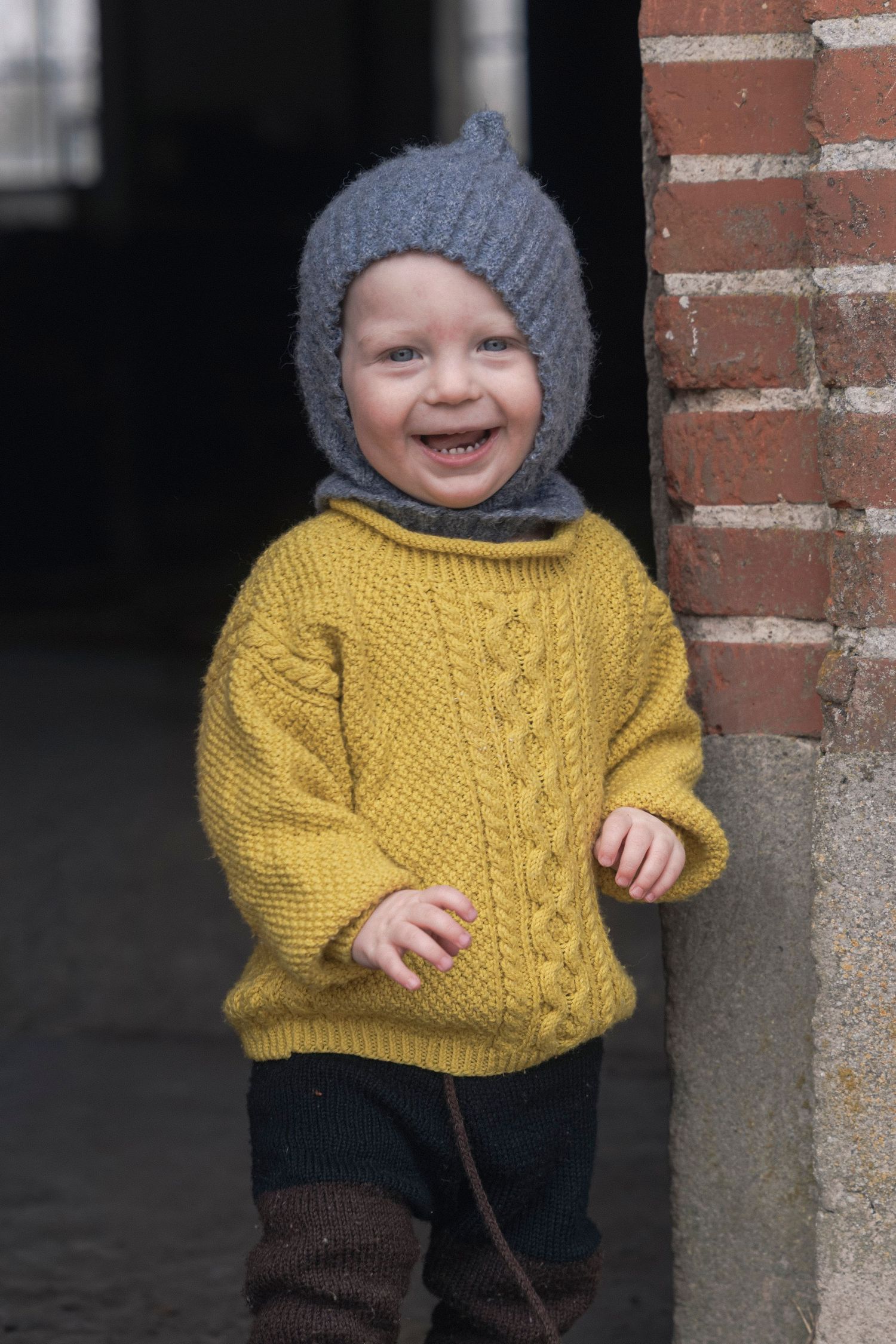 Smiling toddler in yellow cable-knit sweater and gray knit hat standing by brick wall.
