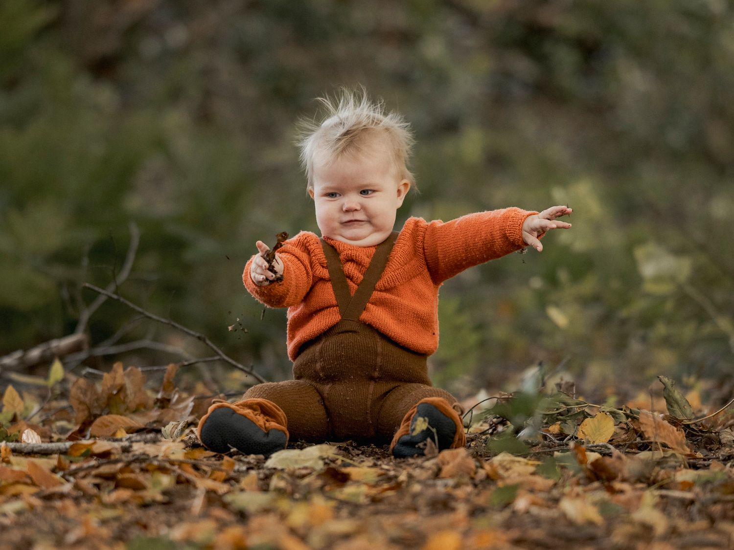 Cheerful baby in orange sweater sitting in autumn leaves outdoors, reaching out with one hand on a fall day.