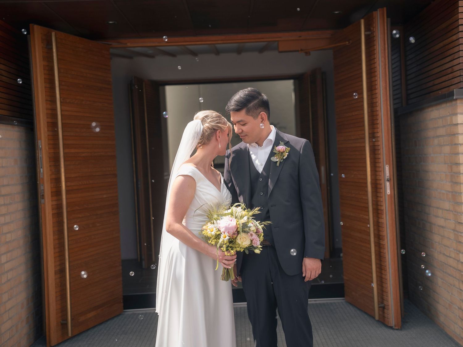 Bride and groom share a romantic moment outside wooden church doors, bride holding pastel bouquet.
