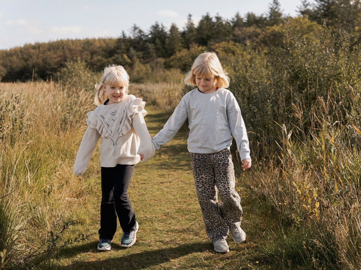 Two young blonde girls holding hands and walking on a grassy path surrounded by tall grass and trees.