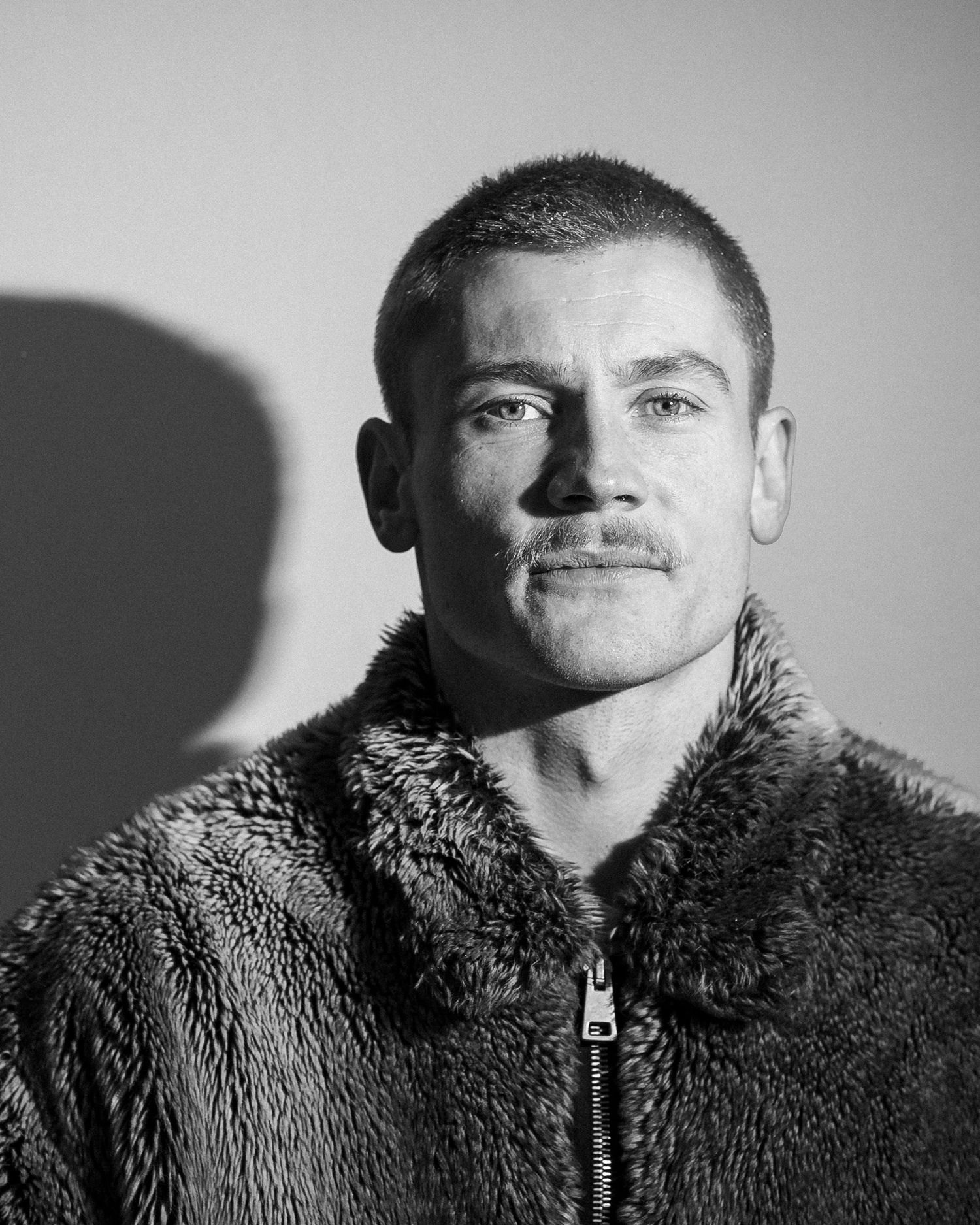 Black and white portrait of a young man with a mustache wearing a textured fur zip-up jacket.