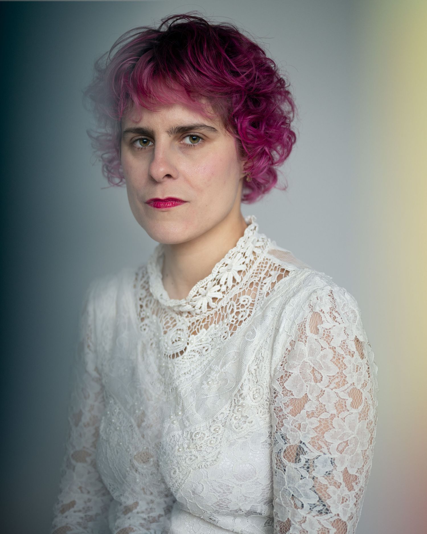 Woman with pink curly hair and red lips wearing a white lace dress poses for a portrait photo.