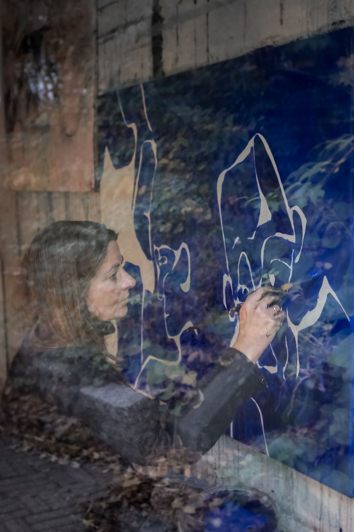 Woman painting a blue mural on a wall, viewed through glass with reflections creating a double exposure effect.