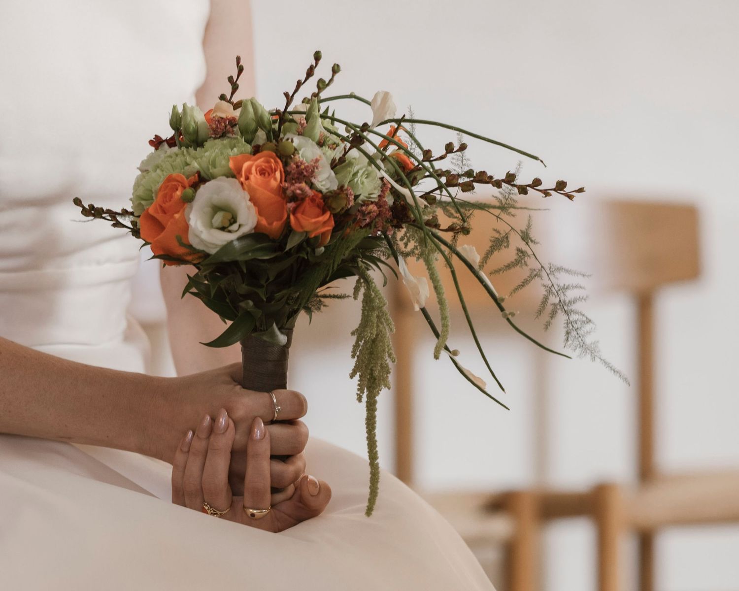 Bride holding an autumn wedding bouquet with orange roses, white flowers, and greenery while wearing a gold ring.