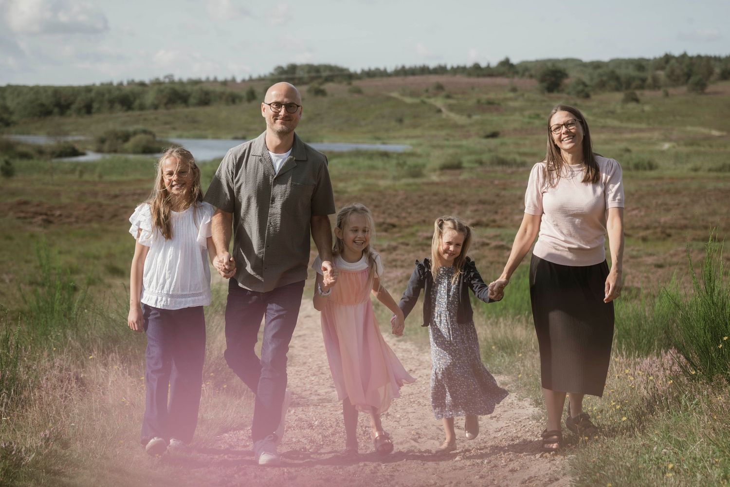 Family of five holding hands on a scenic countryside path surrounded by green fields and pink wildflowers.