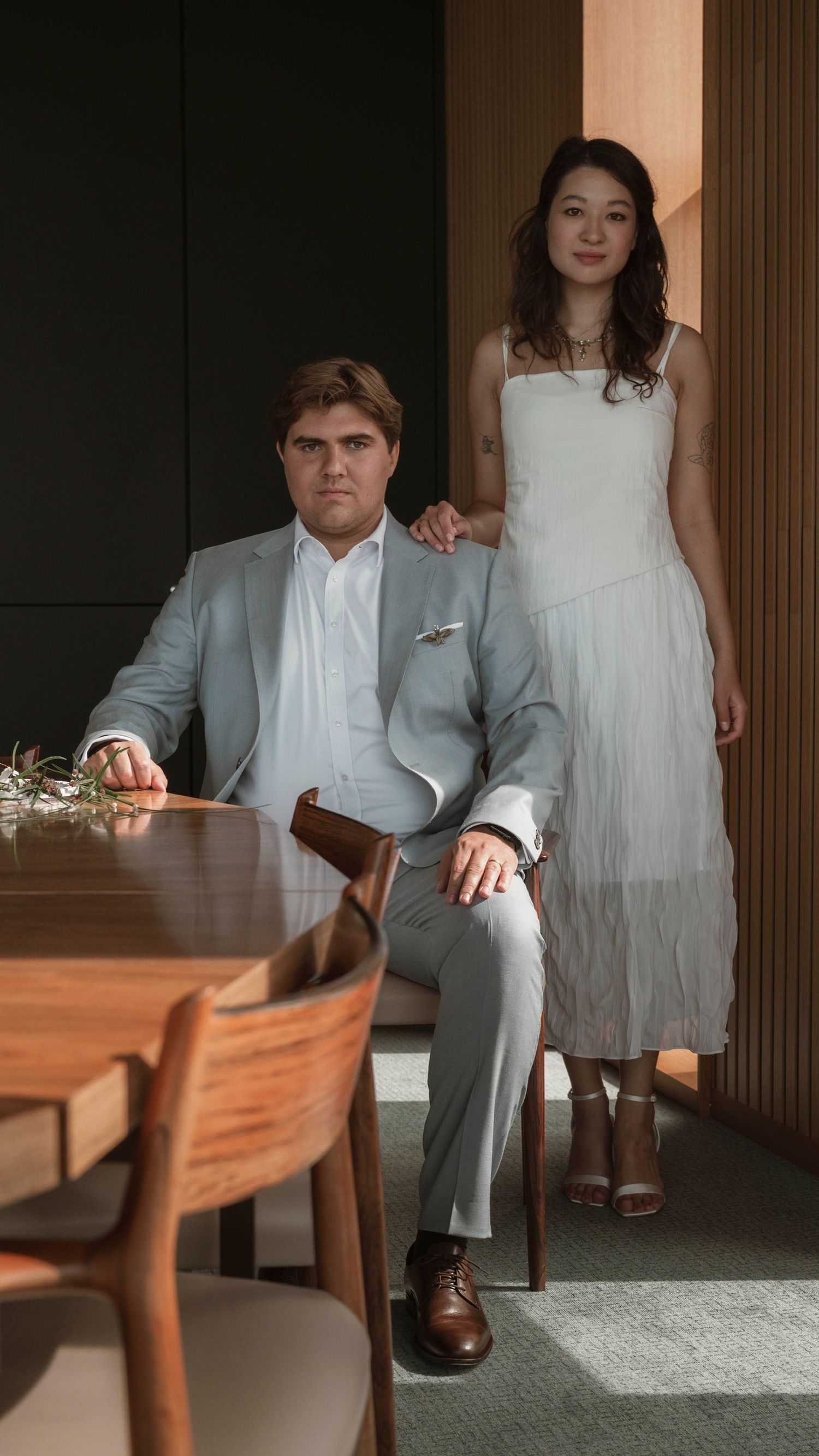 Wedding couple poses in formal attire against dark interior with wooden furniture.