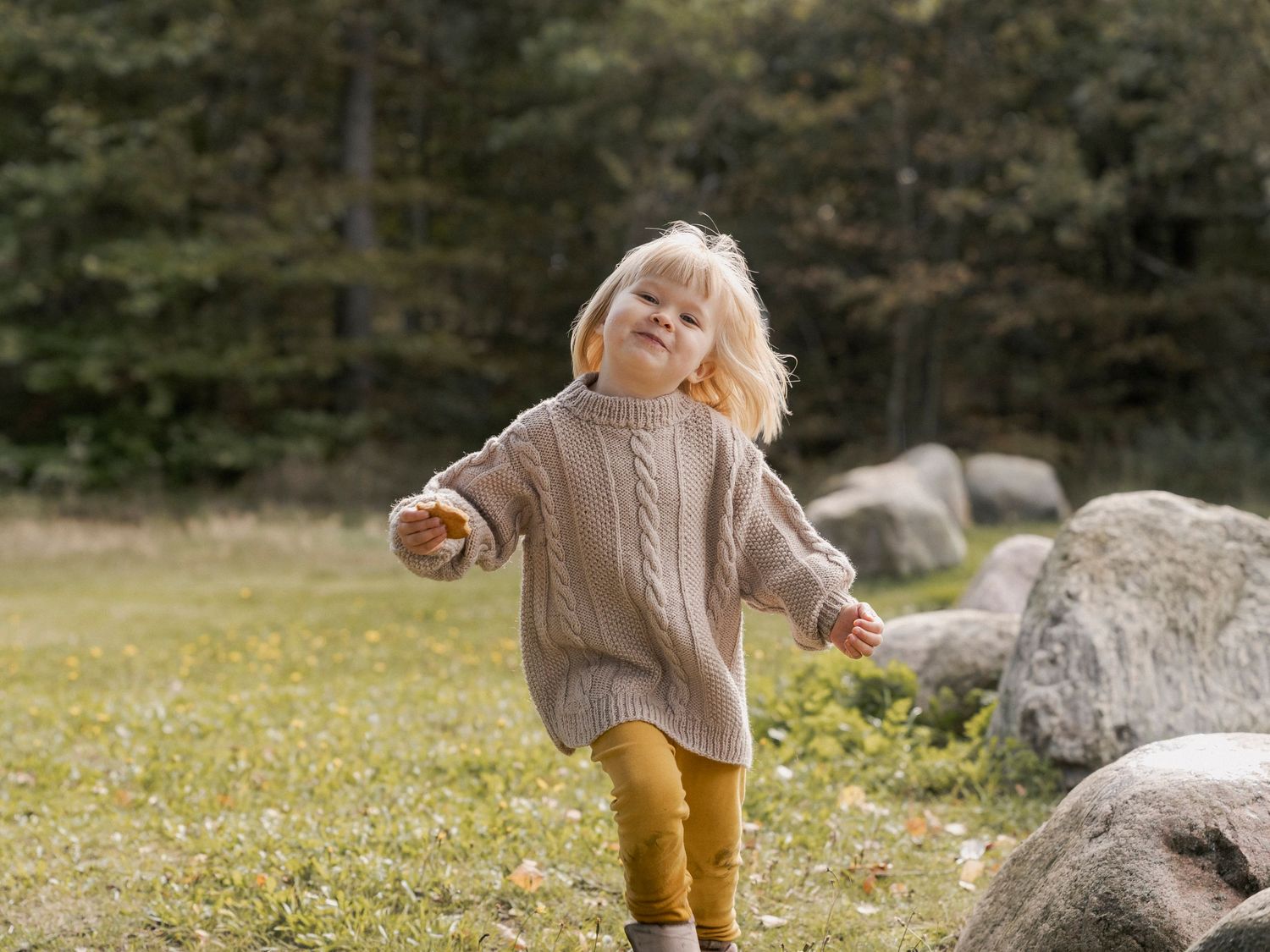 A young blonde toddler girl in a beige knit sweater and mustard pants plays outdoors near rocks and green trees.