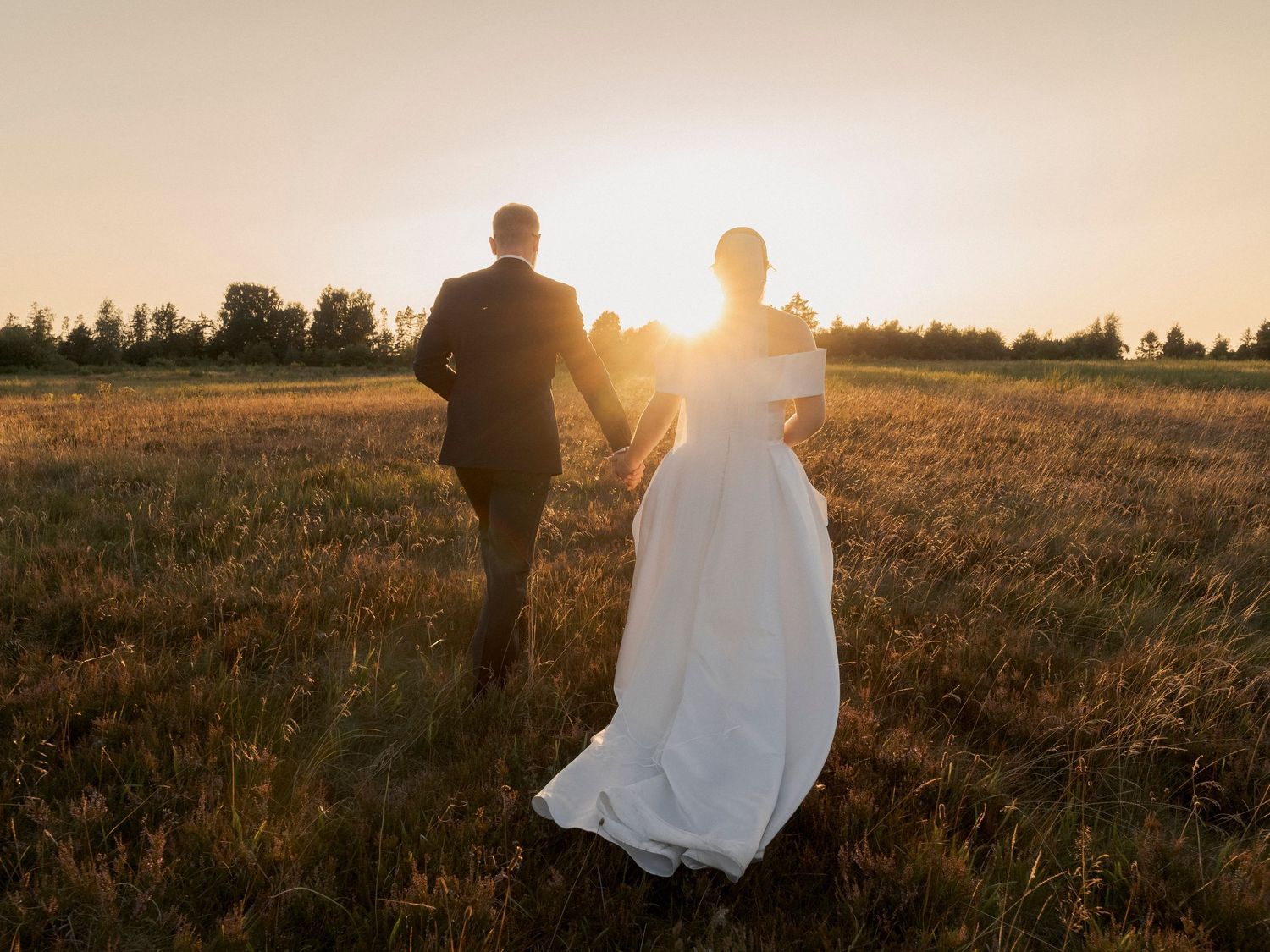 Bride and groom walking hand in hand through golden field at sunset in romantic outdoor wedding photo.