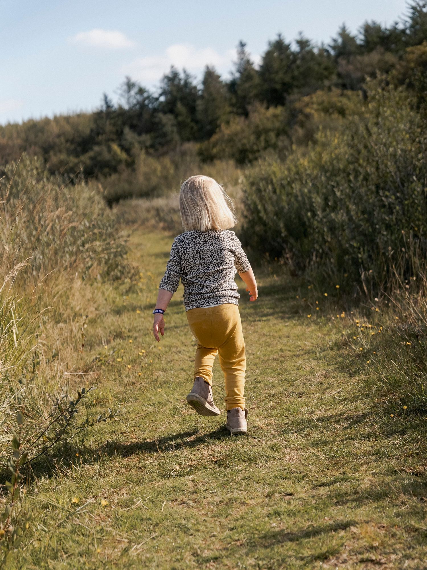 A young child with blonde hair walks along a grassy nature trail surrounded by green shrubs and trees.