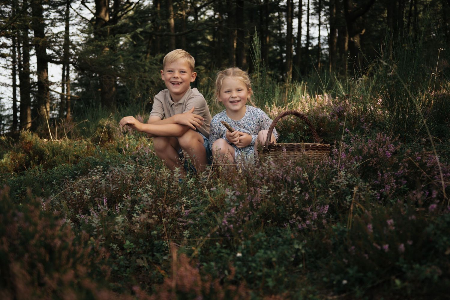 Two smiling children sit together in a lush forest meadow surrounded by wildflowers and tall trees.