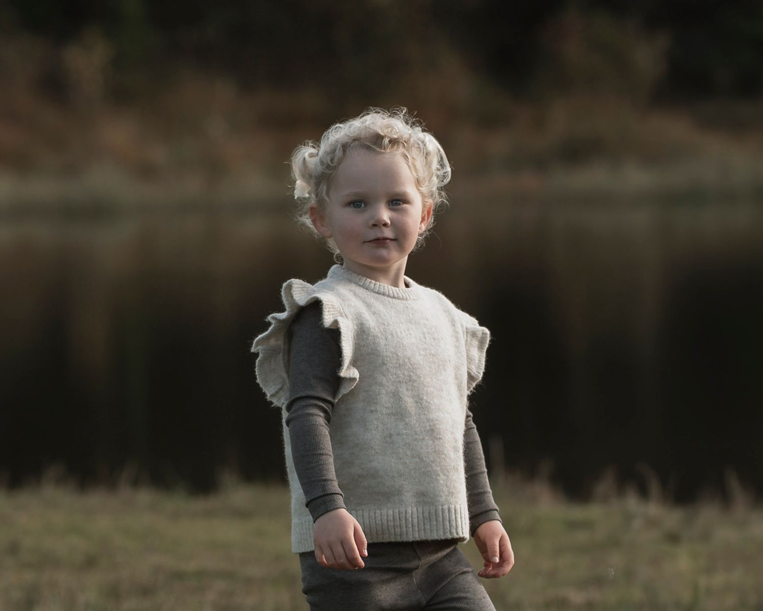 Curly-haired toddler girl wearing a gray ruffle knit vest outdoors near a lake.