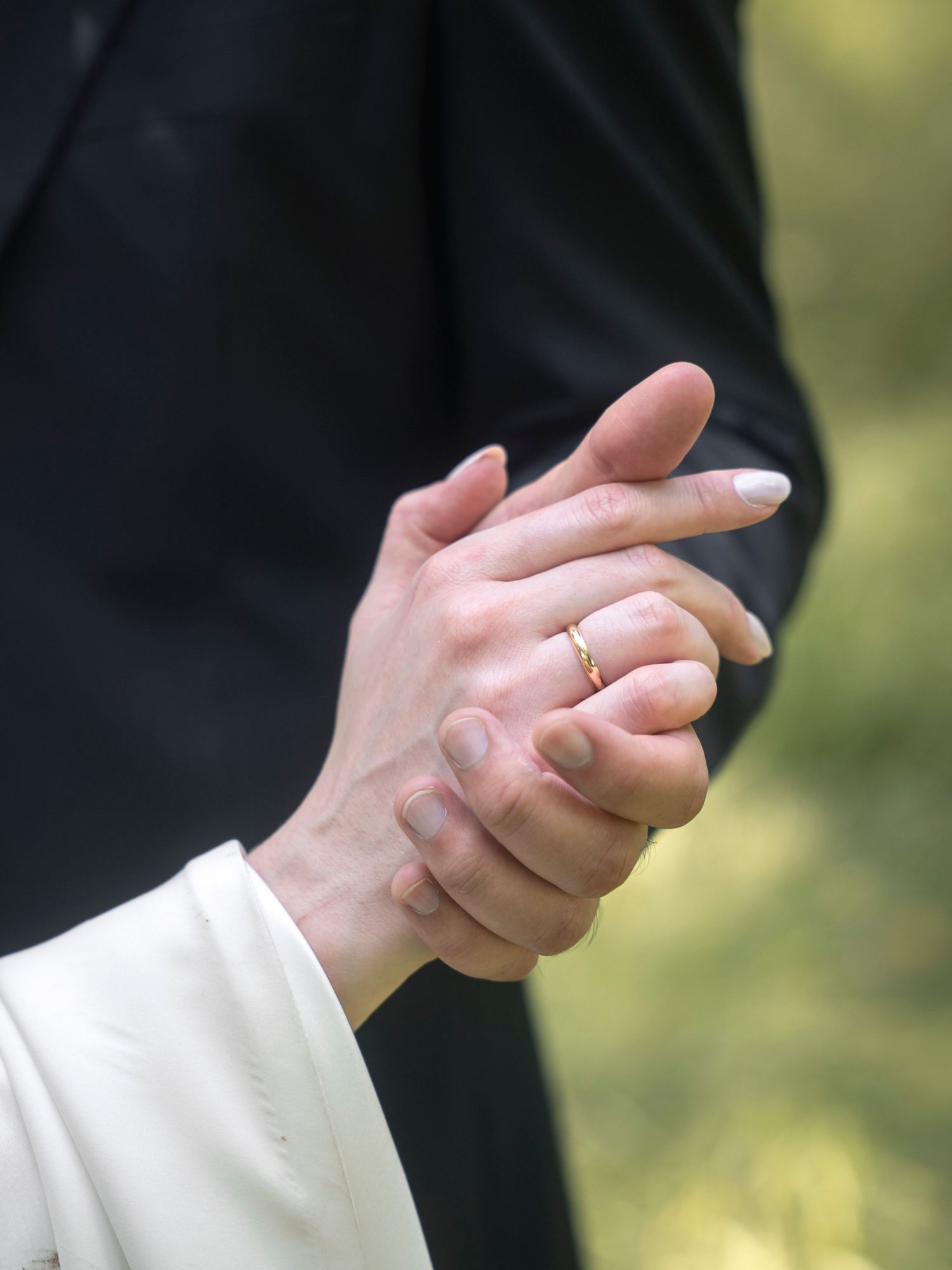 Close-up of couple holding hands, woman wearing gold wedding ring, dressed in white and black attire.