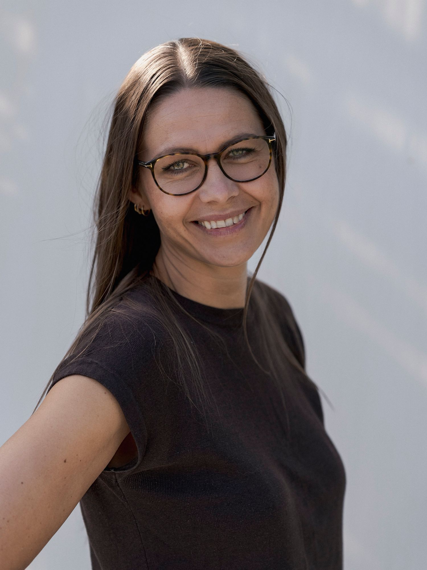 Smiling woman with long brown hair and round glasses wearing a black t-shirt against a light gray background.