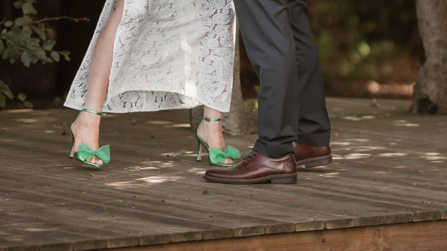 Close-up of a bride in green heels and groom in brown dress shoes standing on a wooden deck at a wedding.