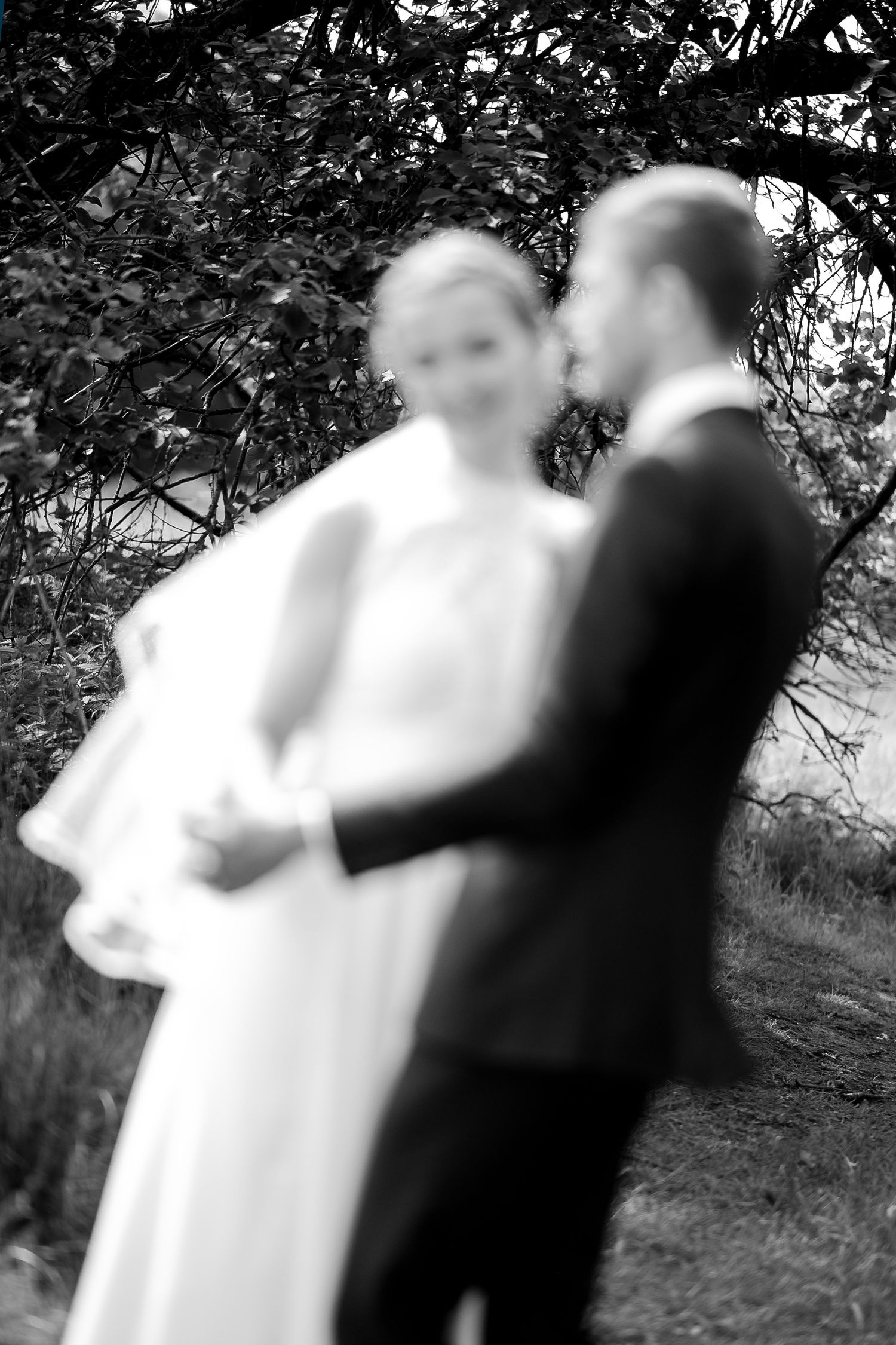 Black and white blurred photo of a bride and groom embracing outdoors under leafy trees on their wedding day.