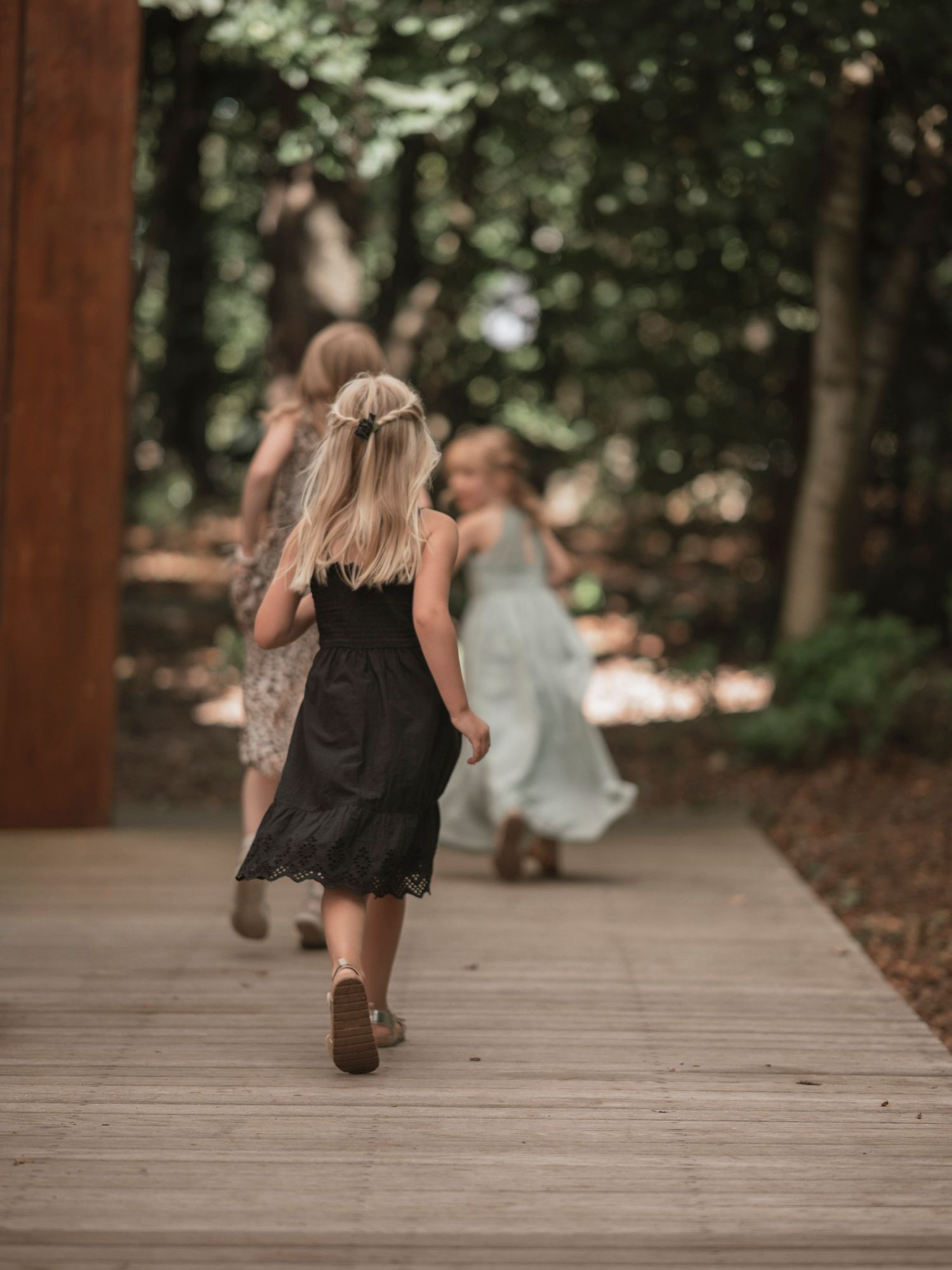 Three young girls in dresses running along a wooden path through a lush green forest.
