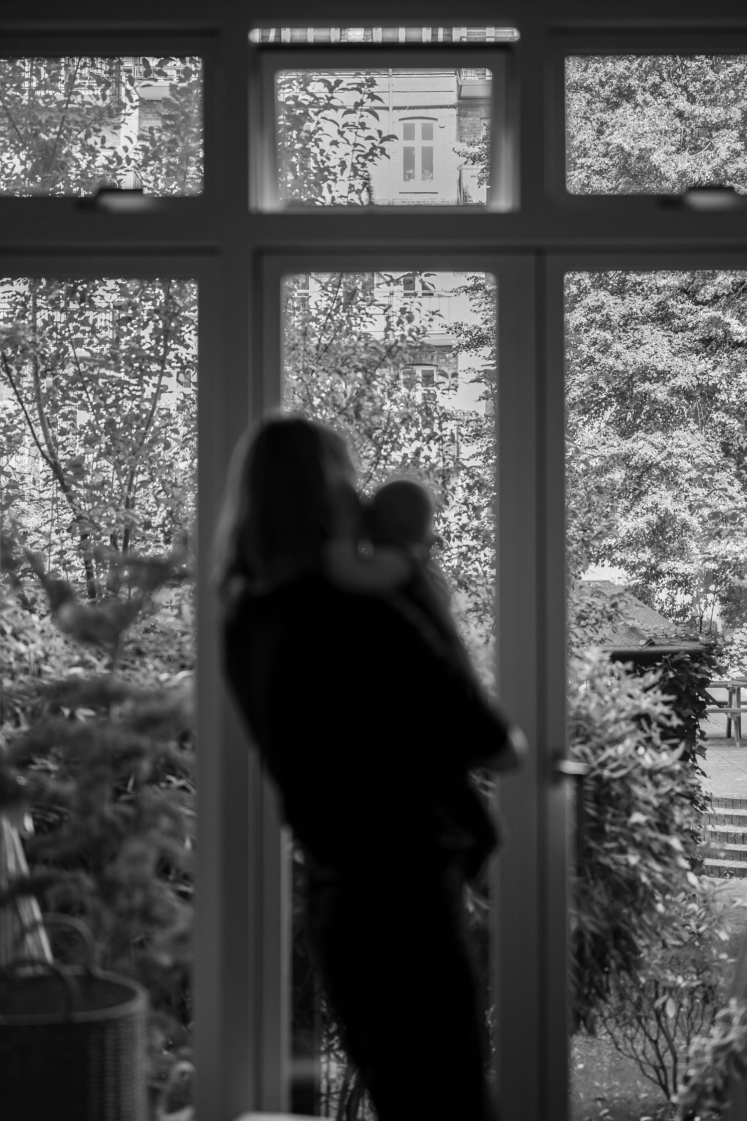 Black and white photo of a mother holding her baby, silhouetted against a large window with trees outside.