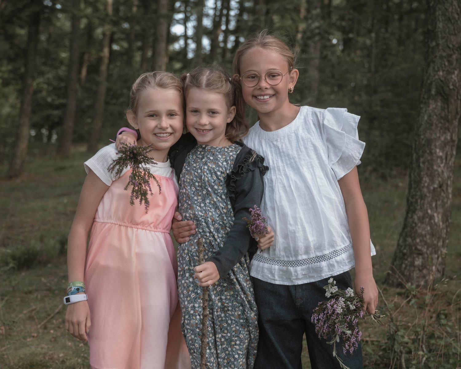 Three young girls smiling and hugging outdoors in a forest setting, wearing elegant dresses for a special occasion.