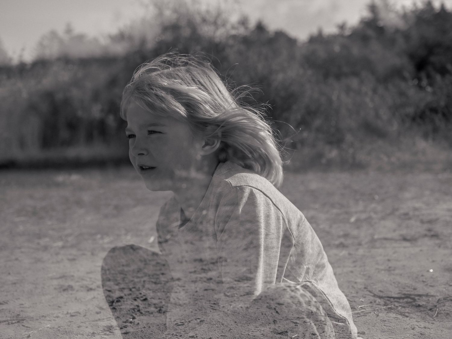 Black and white photo of a young child with windswept hair playing in the sand outdoors.