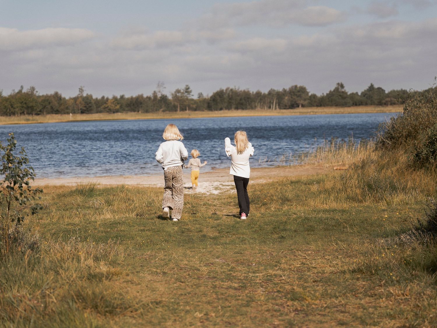 Two adults and a child walk toward a serene lake surrounded by lush greenery under a partly cloudy sky.