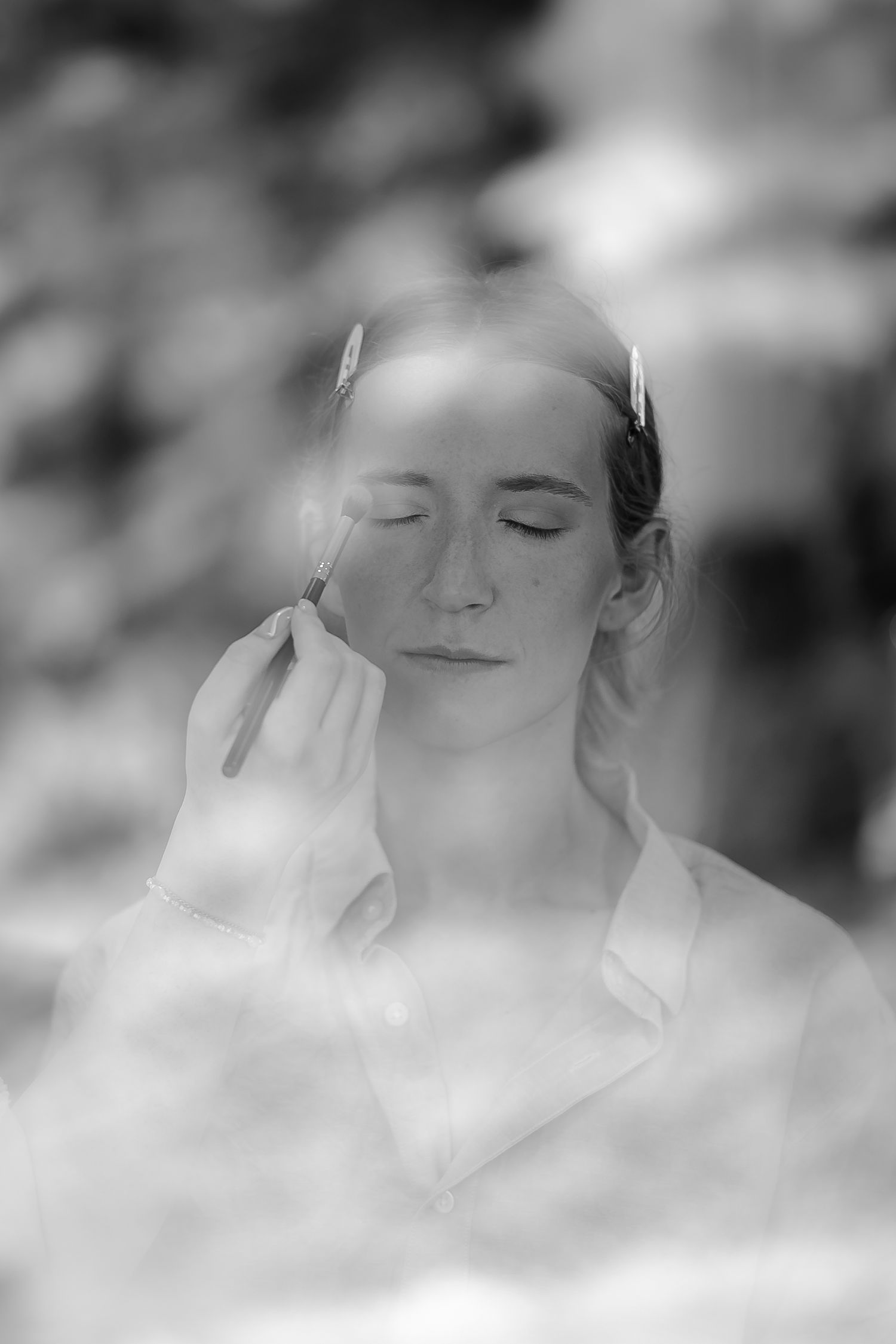 Black and white portrait of a woman with closed eyes having makeup applied outdoors.