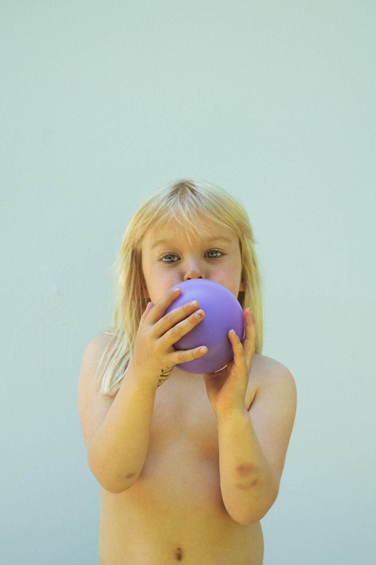 Young blonde child holding a purple ball up to their face against a light blue background.