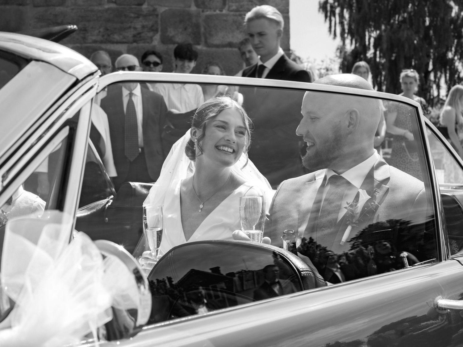 Black and white photo of a smiling bride and groom sitting in a classic wedding car holding champagne glasses.