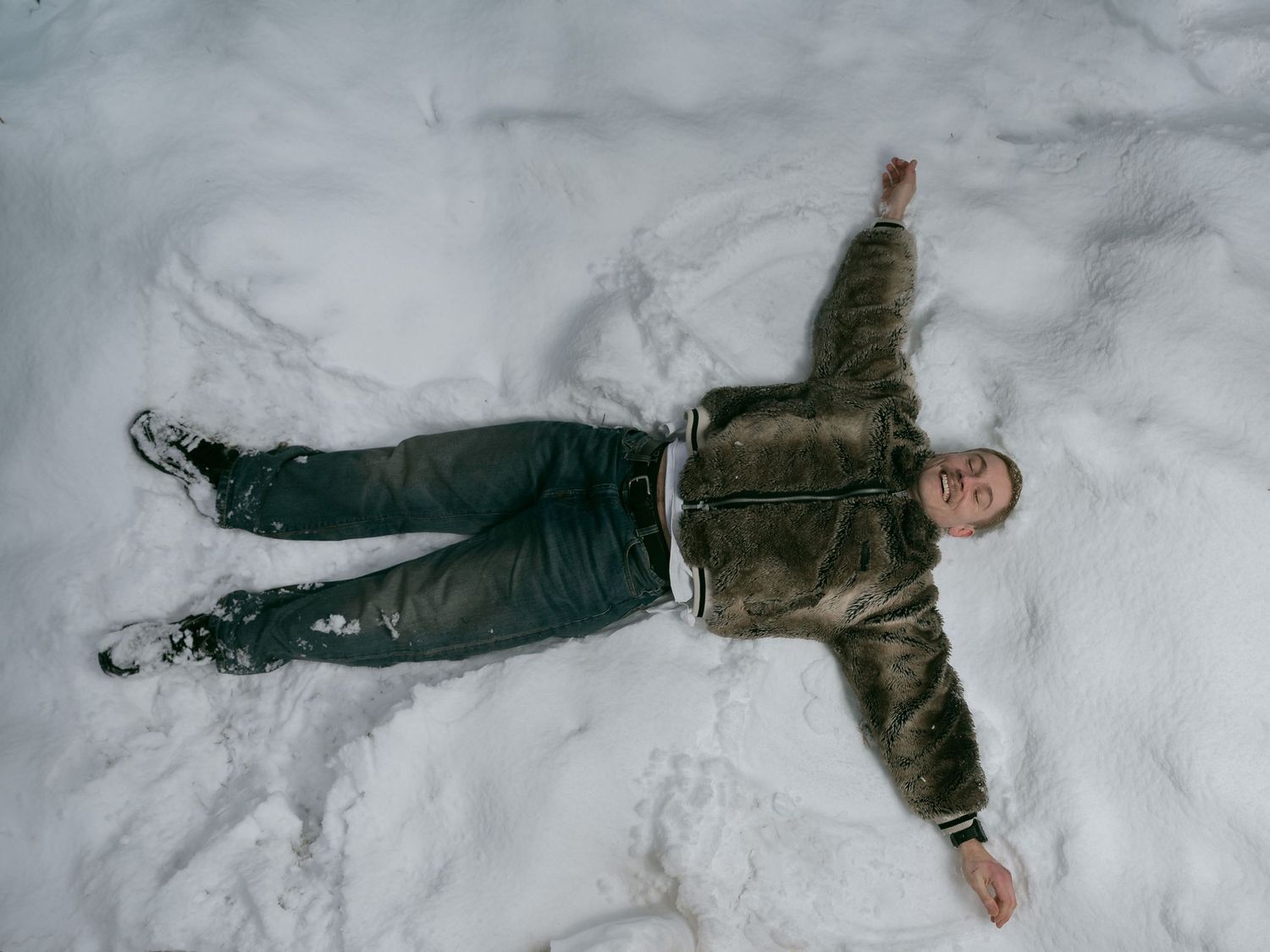 Man lying in snow making a snow angel, wearing a brown jacket and dark pants outdoors in winter.