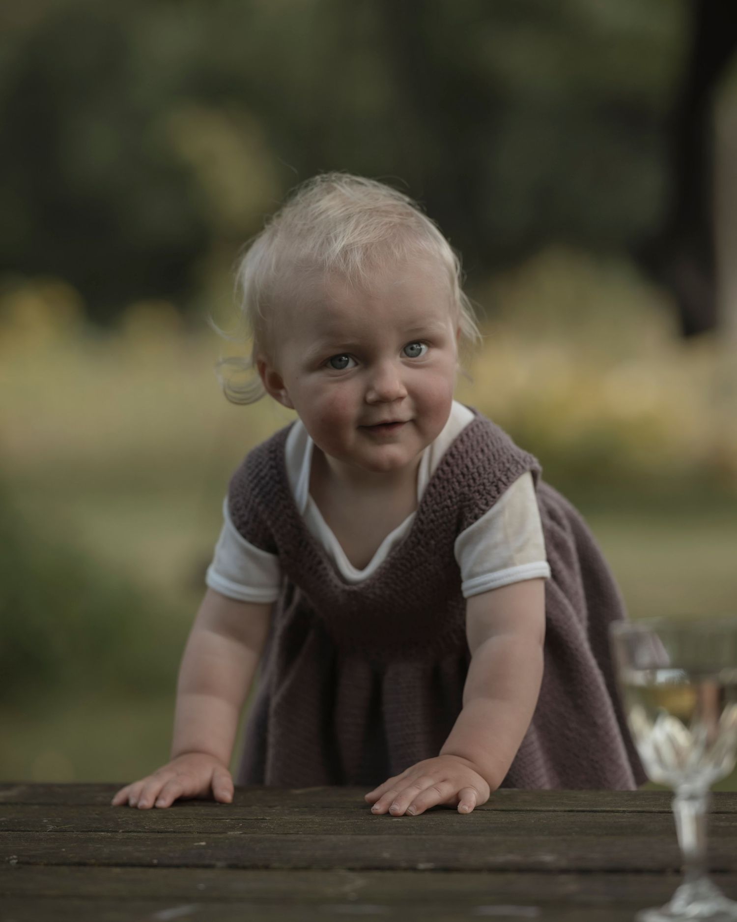 Smiling baby in a brown knit vest dress leaning on a wooden table outdoors in a green garden setting.