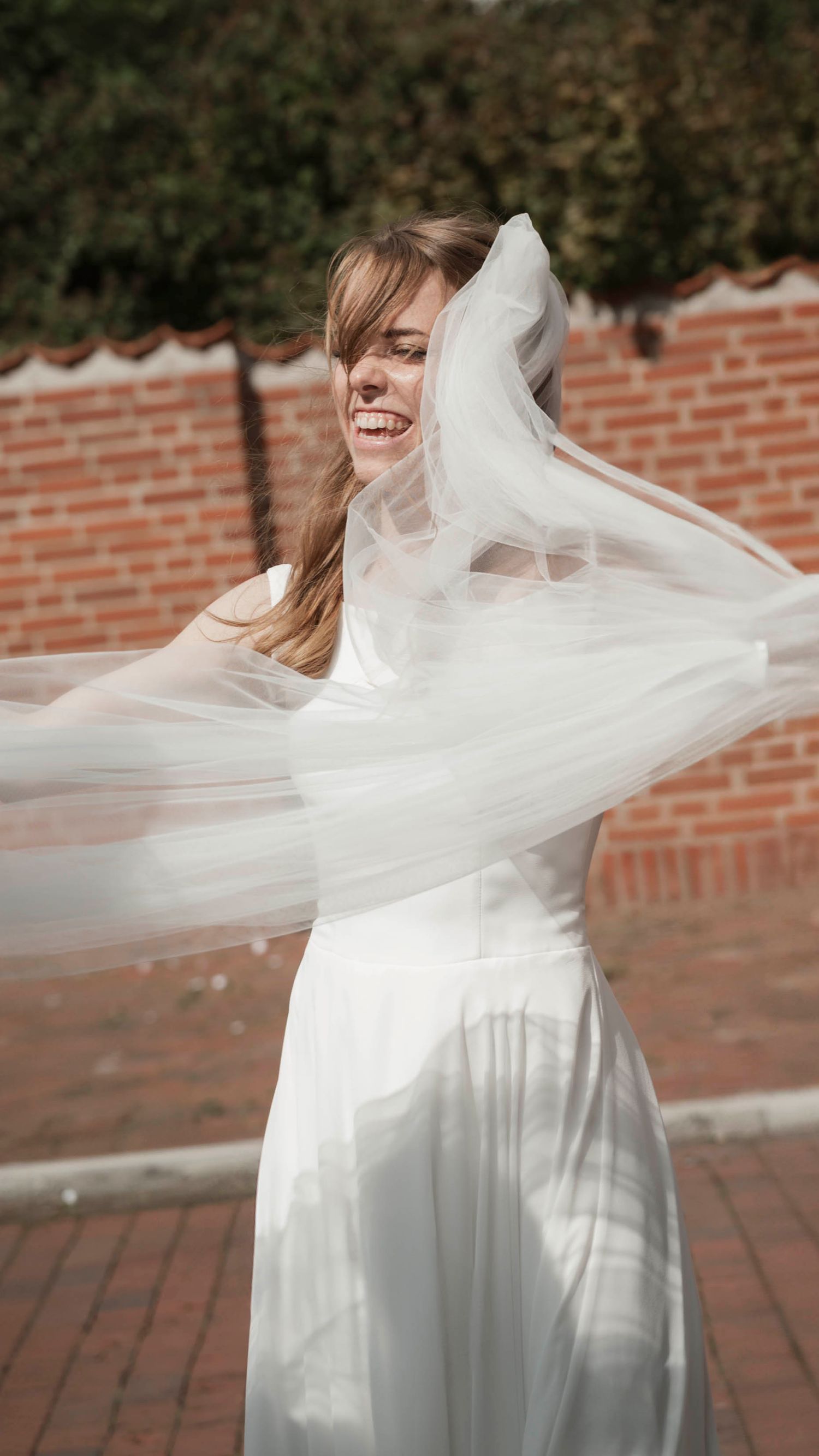 Joyful bride in white gown twirling with flowing veil outdoors near brick wall on her wedding day.
