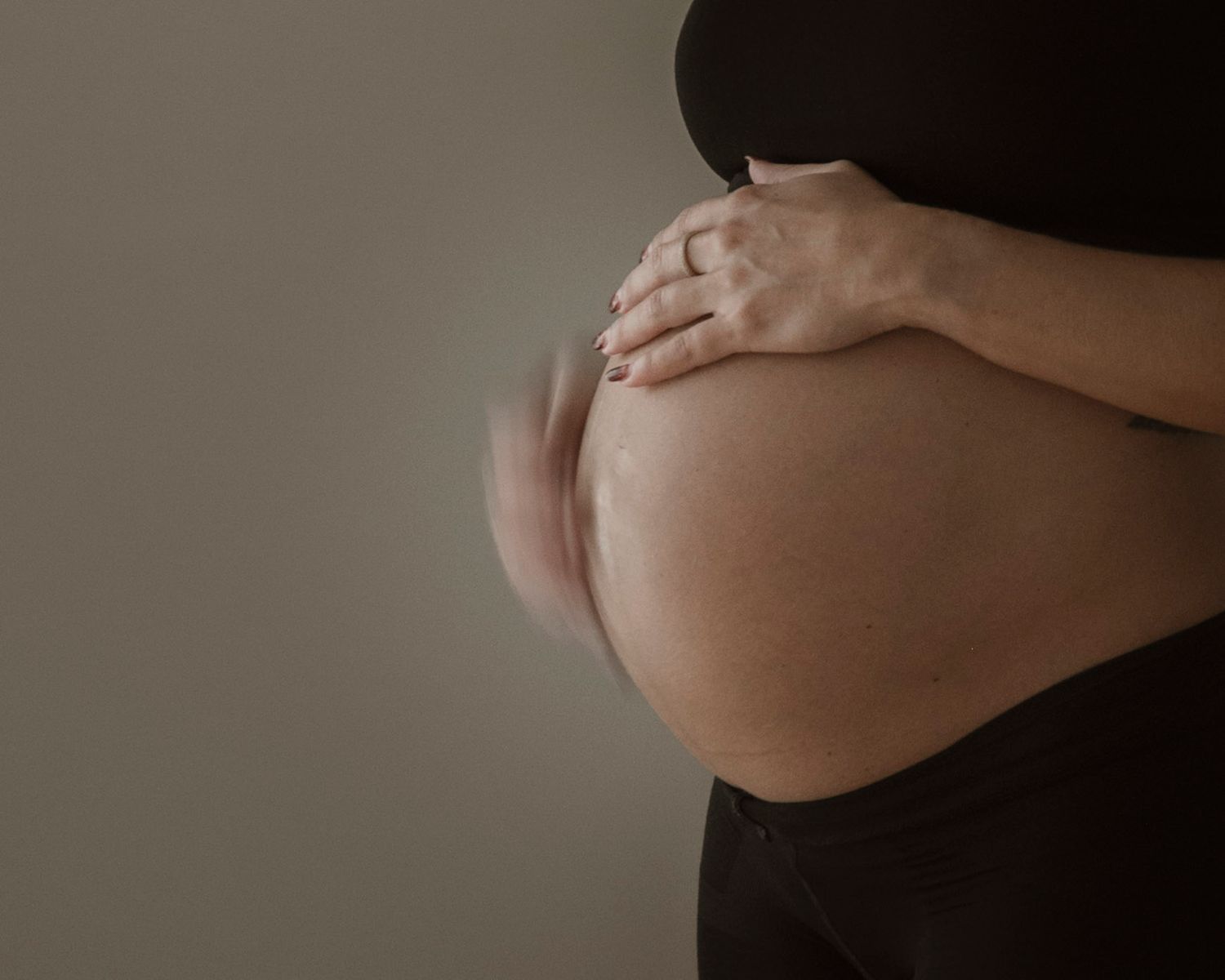 Close-up of a pregnant woman cradling her bare baby bump with both hands against a neutral background.