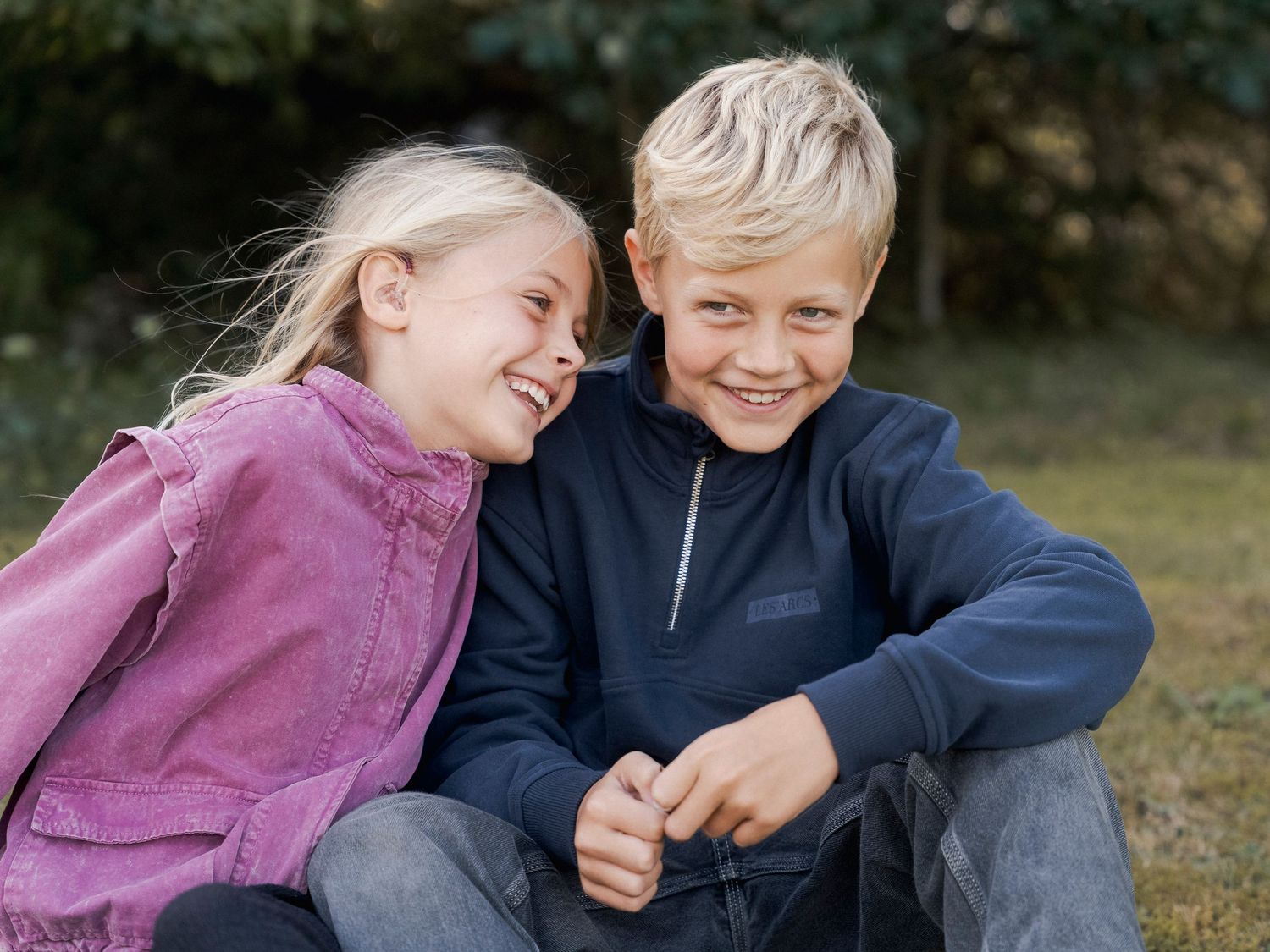 Two happy children, a blonde girl in pink and a boy in navy, sit together smiling outdoors on grass.