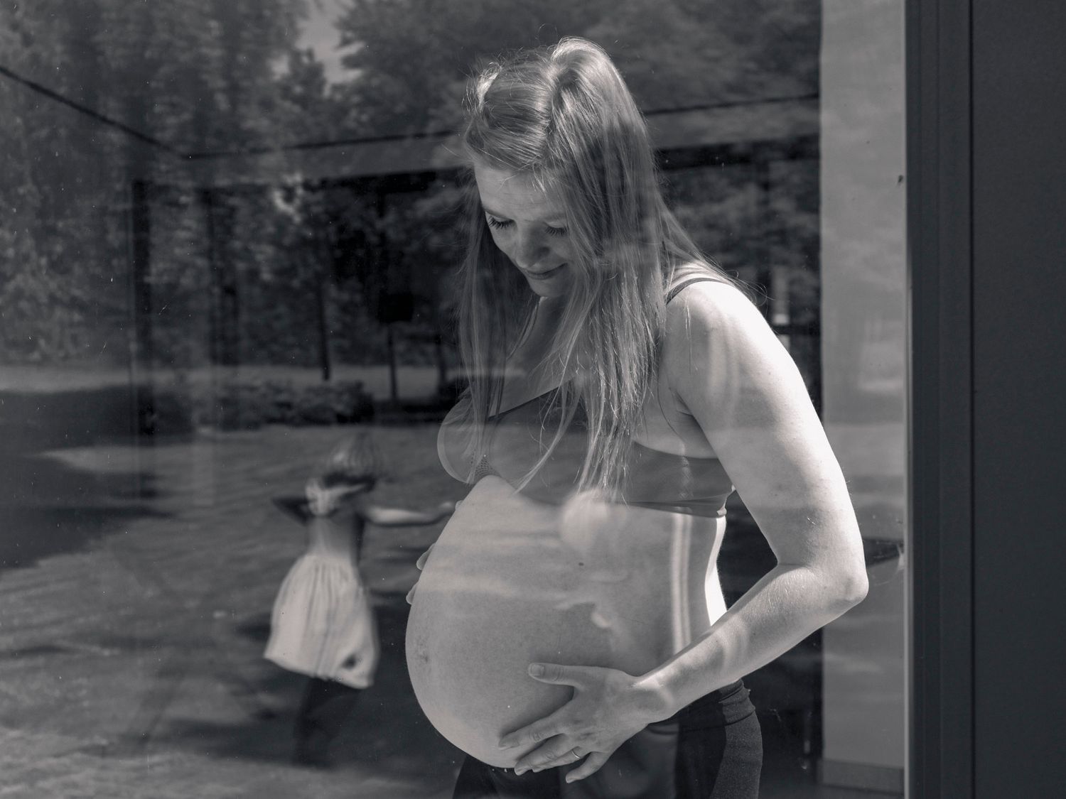 Black and white photo of a pregnant woman cradling her belly, standing by a window with a child visible outside.