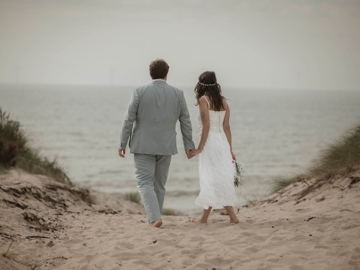 A couple in wedding attire walks down a sandy beach path towards the ocean on an overcast day.