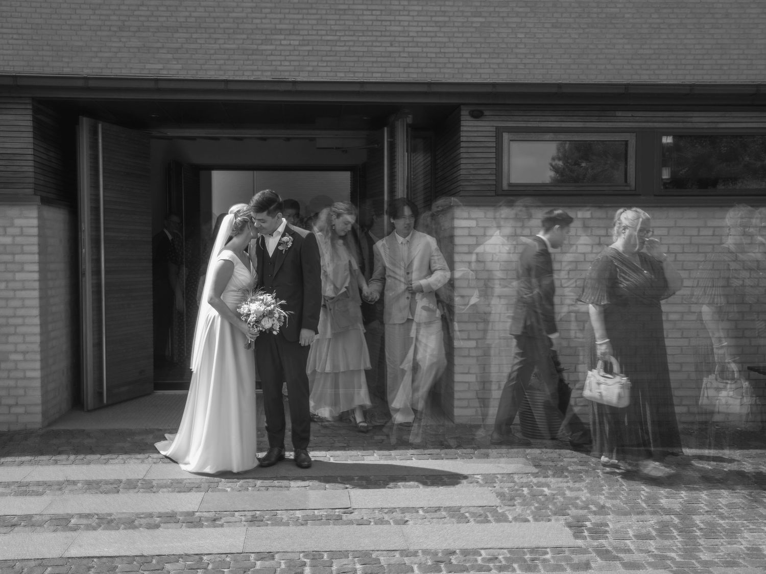 Black and white photo of a bride and groom outside a brick church with wedding guests queuing nearby.