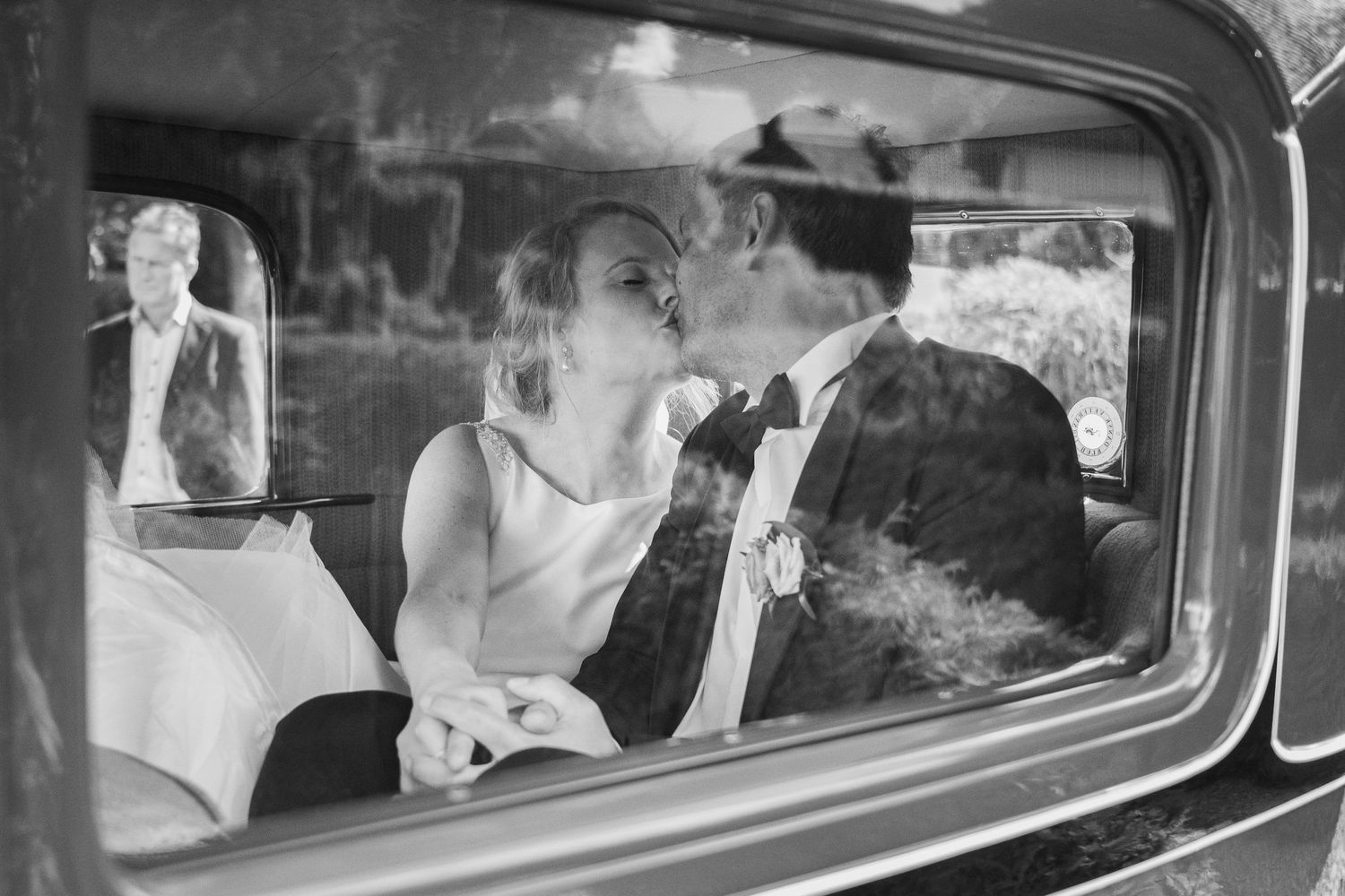 Black and white photo of a bride and groom sharing a kiss inside a vintage car on their wedding day.