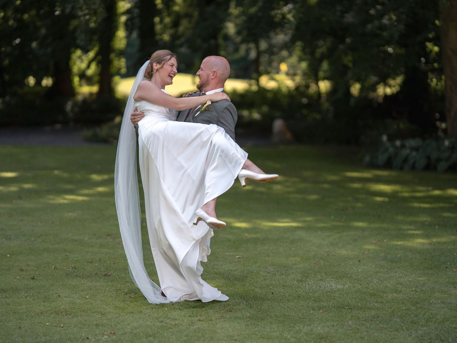 Groom lifting bride in white wedding dress and veil on lush green lawn surrounded by trees.