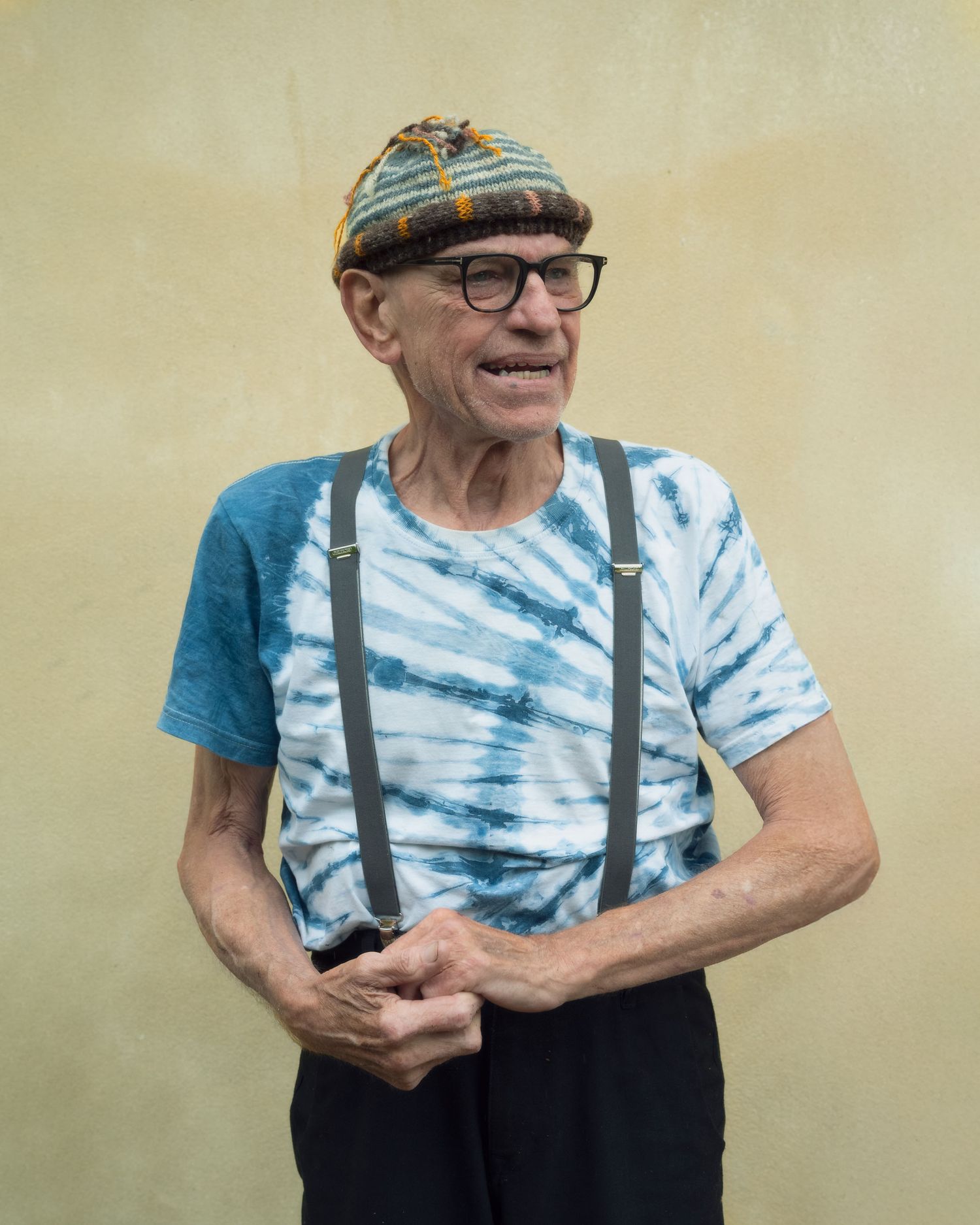 Elderly man in tie-dye shirt, suspenders, and knit cap smiling outdoors against a yellow wall.