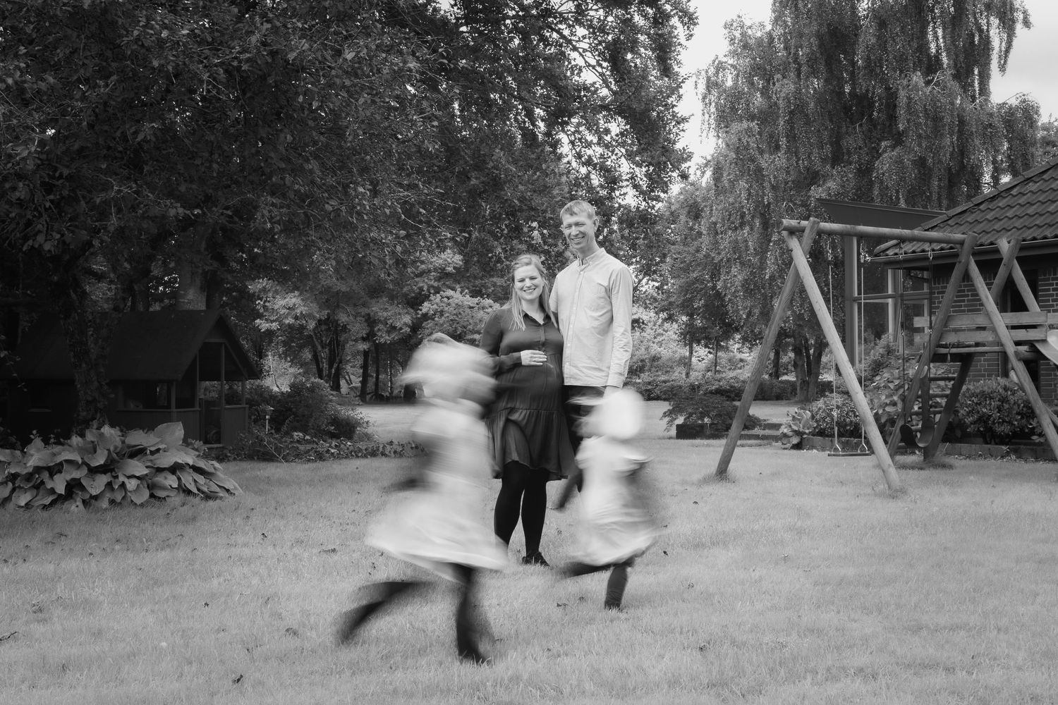 Black and white photo of couple standing in garden while two children spin and play near a wooden swing set.