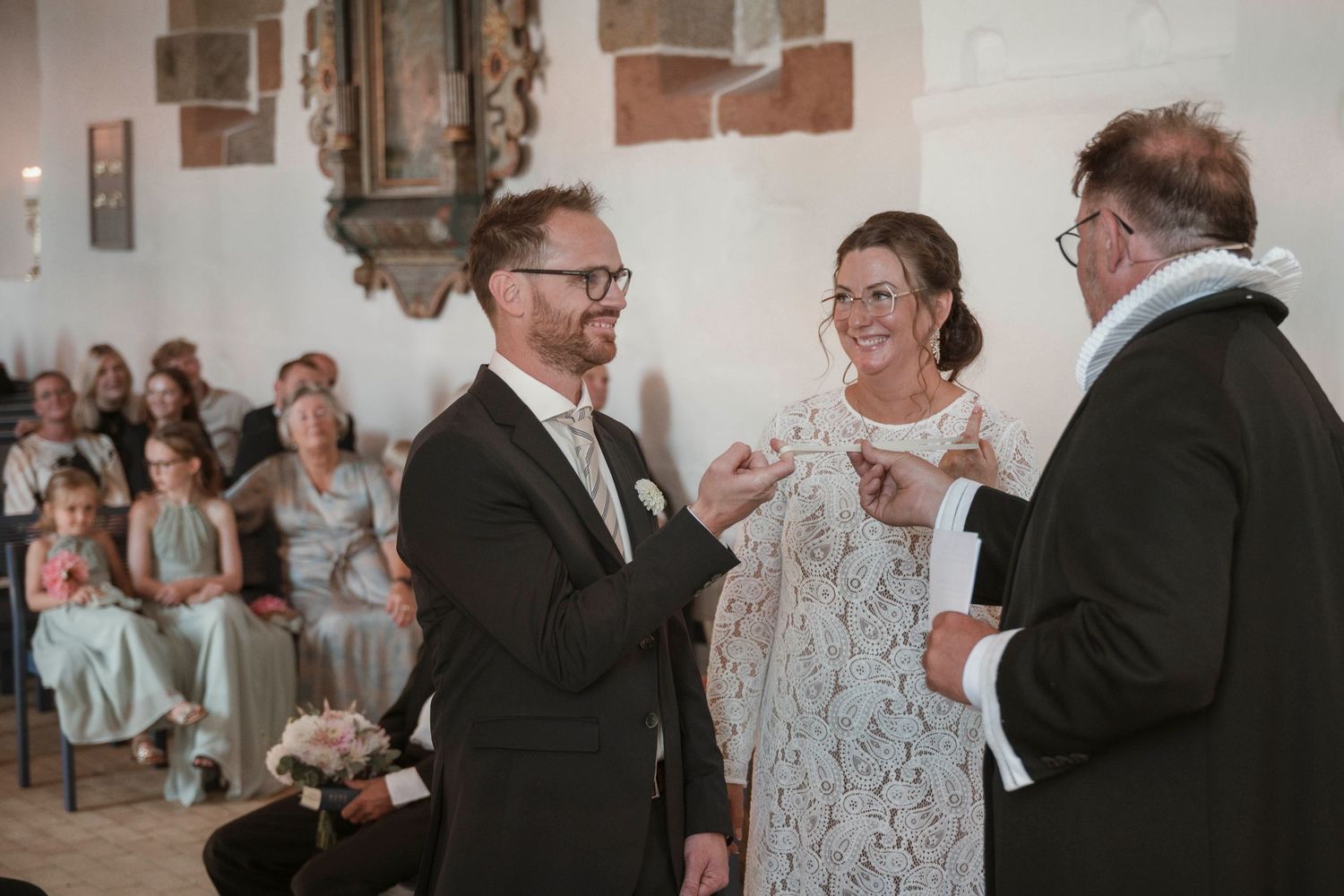 Couple exchanging wedding rings during a church ceremony, smiling joyfully, with guests seated in the background.