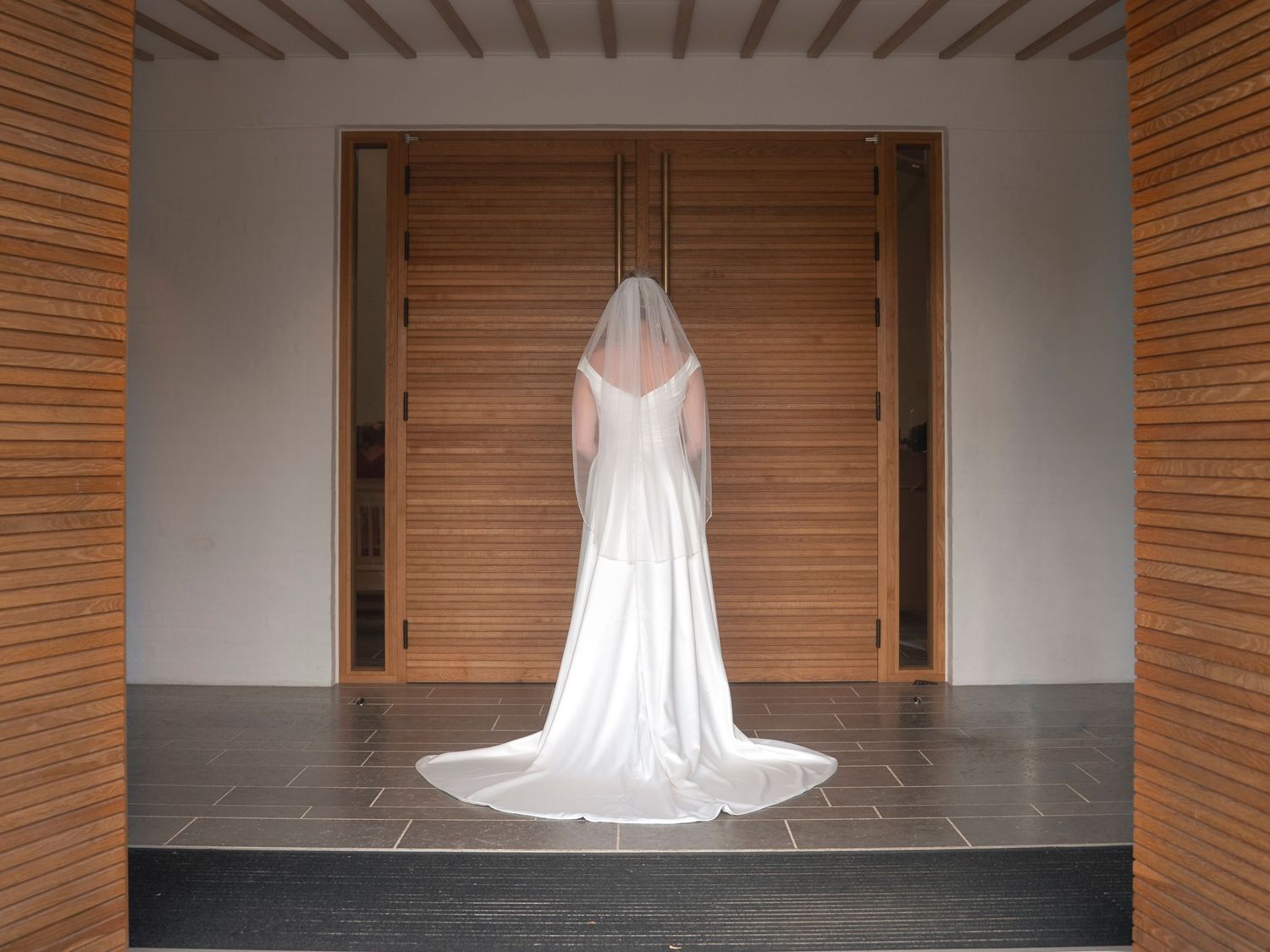 Bride in white wedding gown and veil standing before large wooden doors in elegant venue.