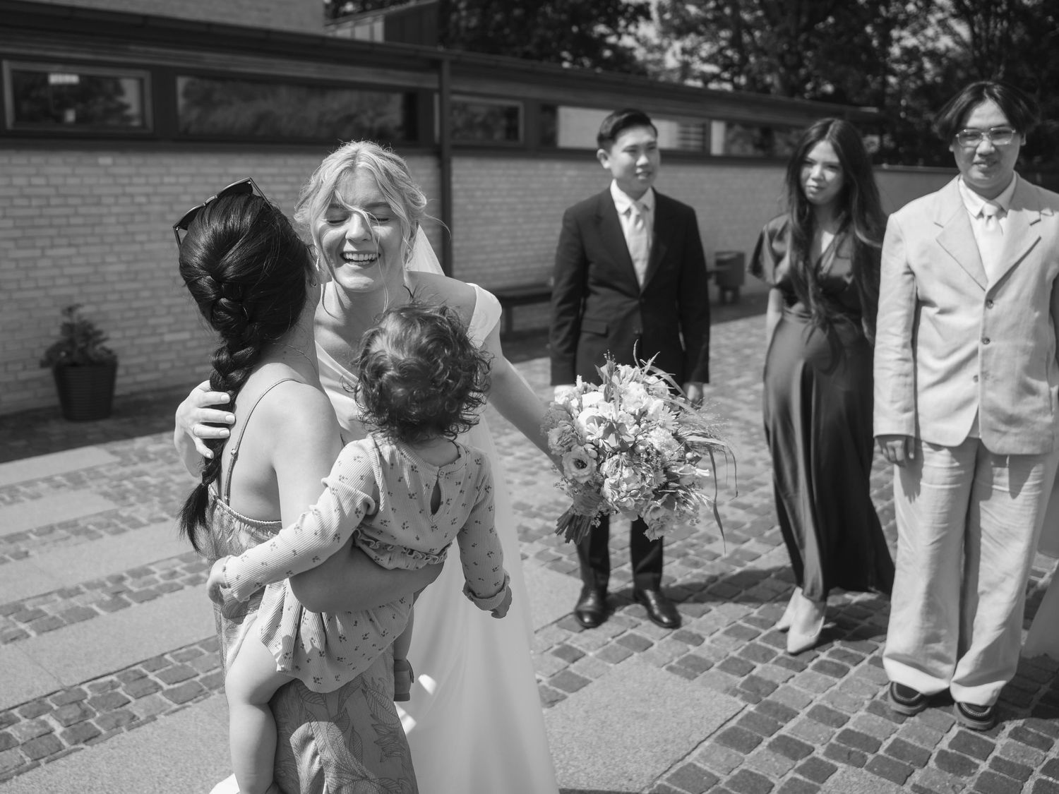 Black and white wedding photo of a bride hugging a guest holding a child, with wedding guests standing nearby outdoors.