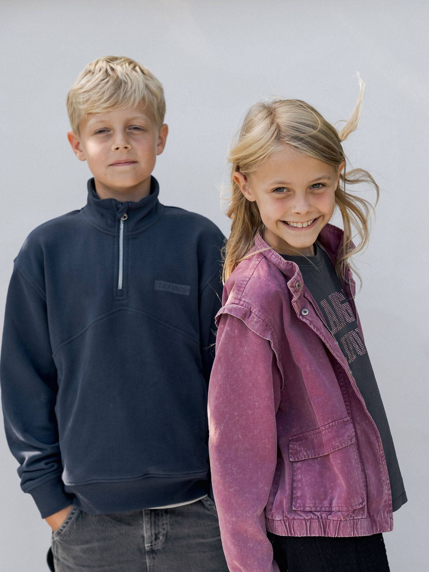 Two kids posing outdoors, boy in navy half-zip sweatshirt and girl in pink denim jacket smiling.
