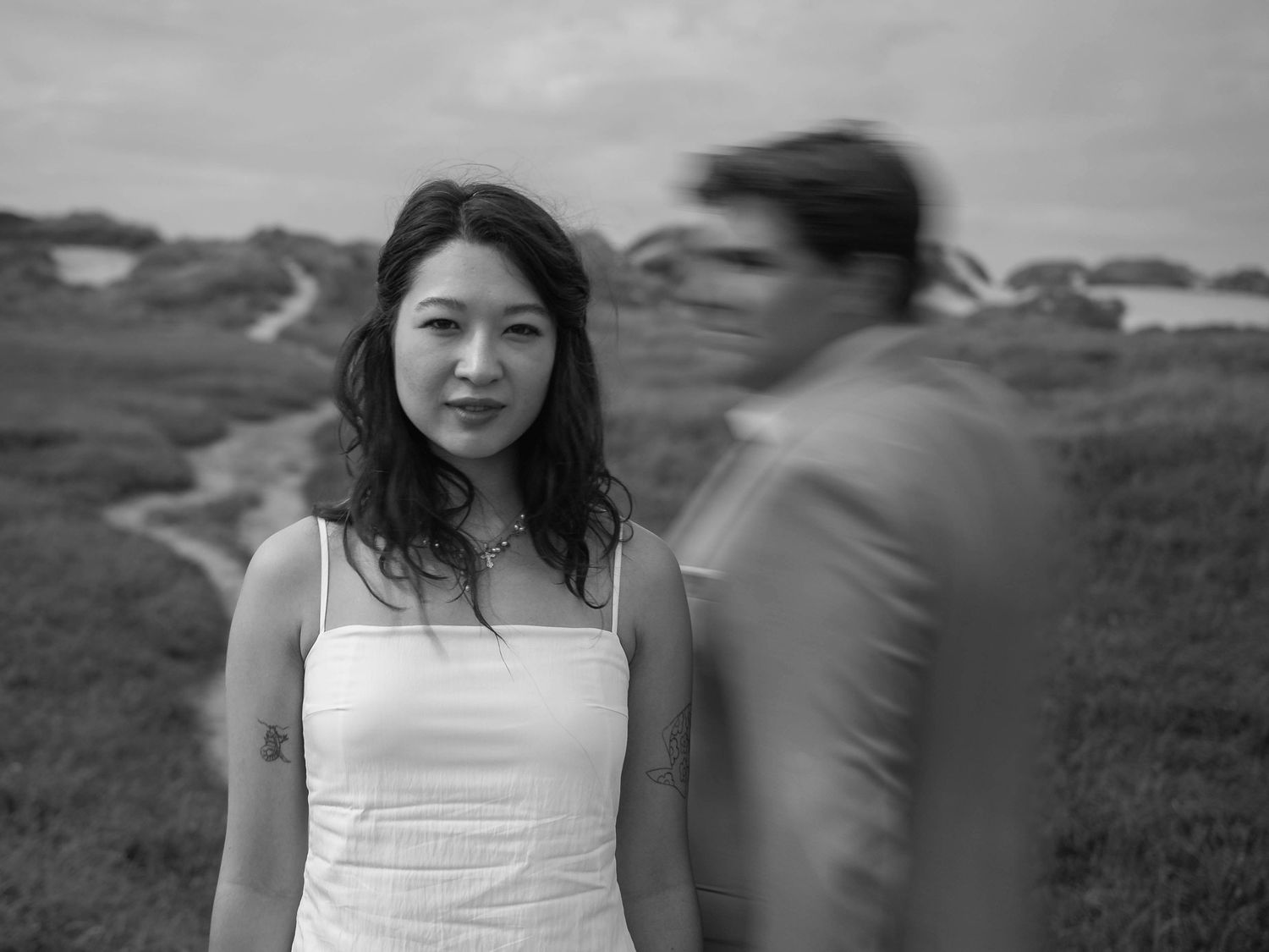 Black and white photo of a woman in a white dress standing outdoors with a blurred man in the foreground.