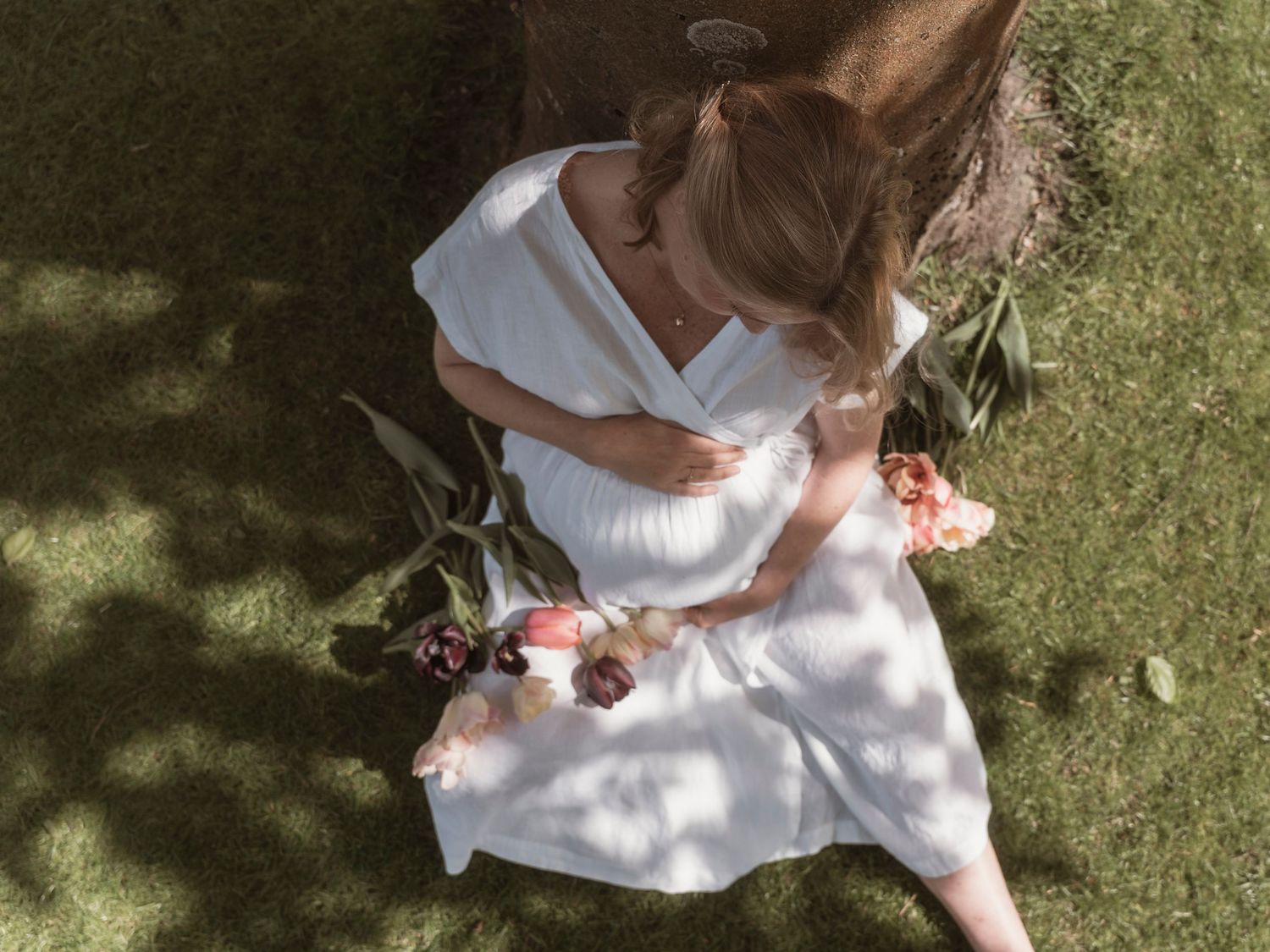 Aerial view of a pregnant woman in a white dress sitting on grass, cradling her belly while holding a bouquet of flowers.
