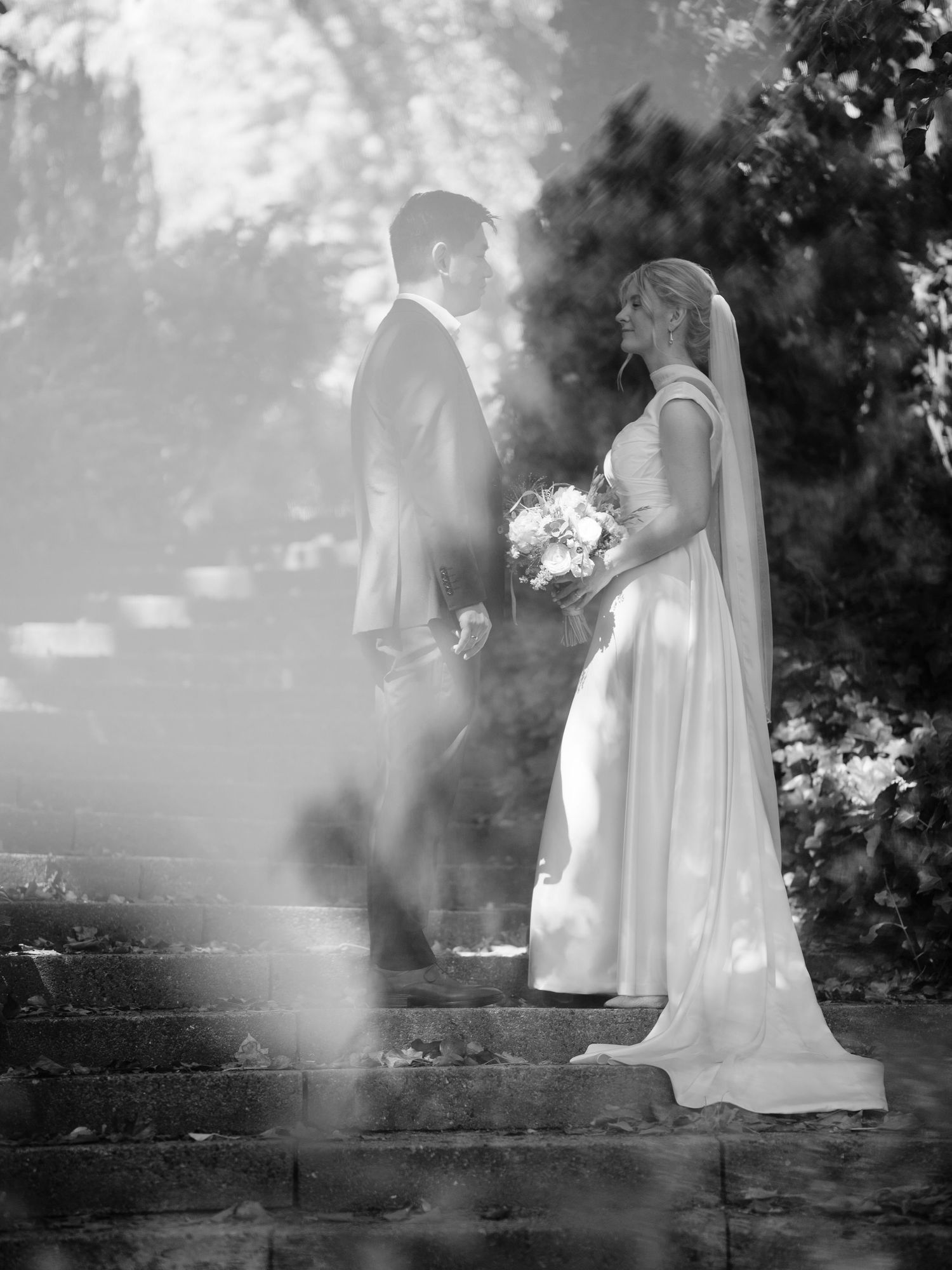 Black and white wedding photo of bride holding bouquet and groom standing on stone steps outdoors.