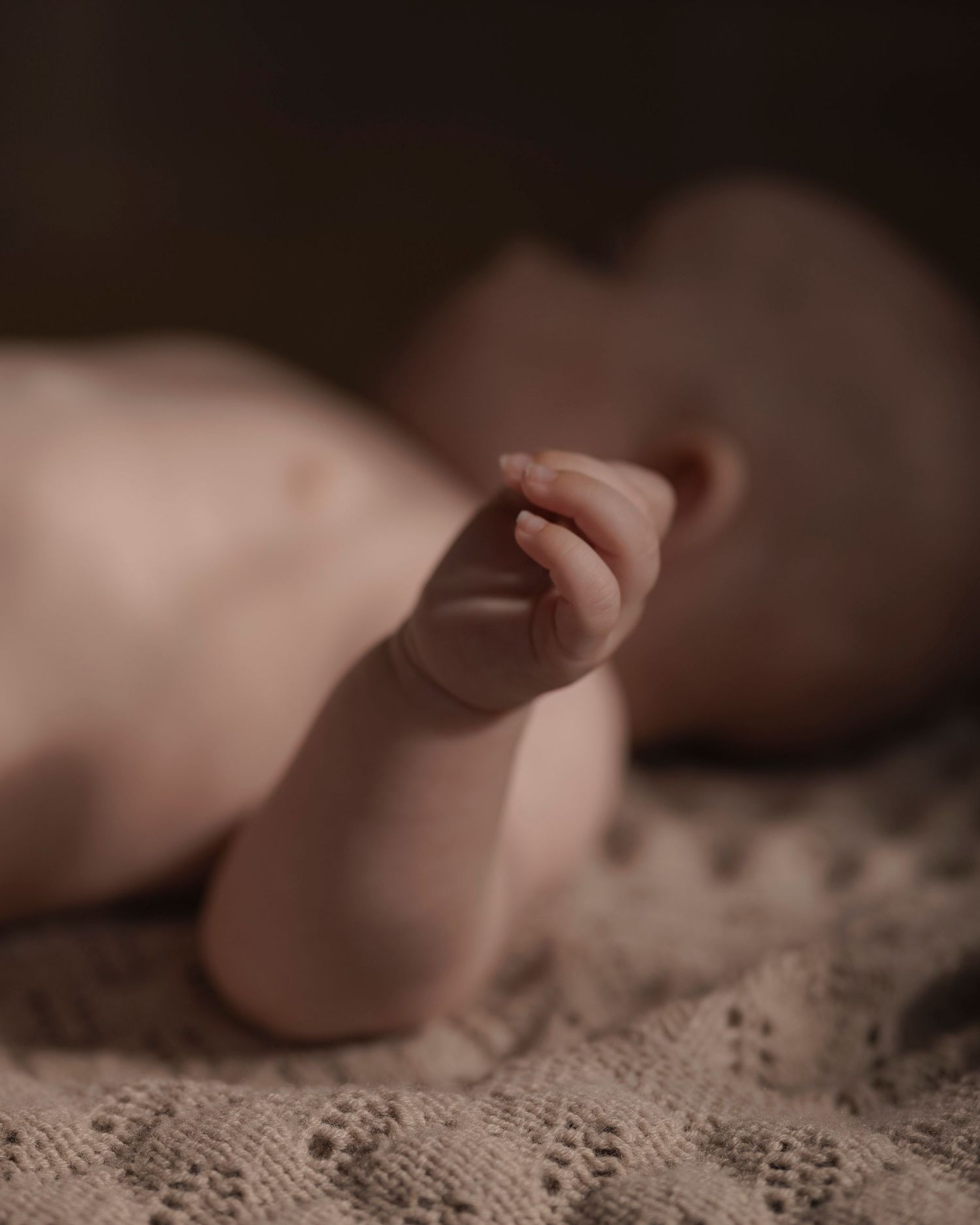 Close-up of a newborn baby's tiny hand raised up while lying on a soft knit blanket in warm, dim light.