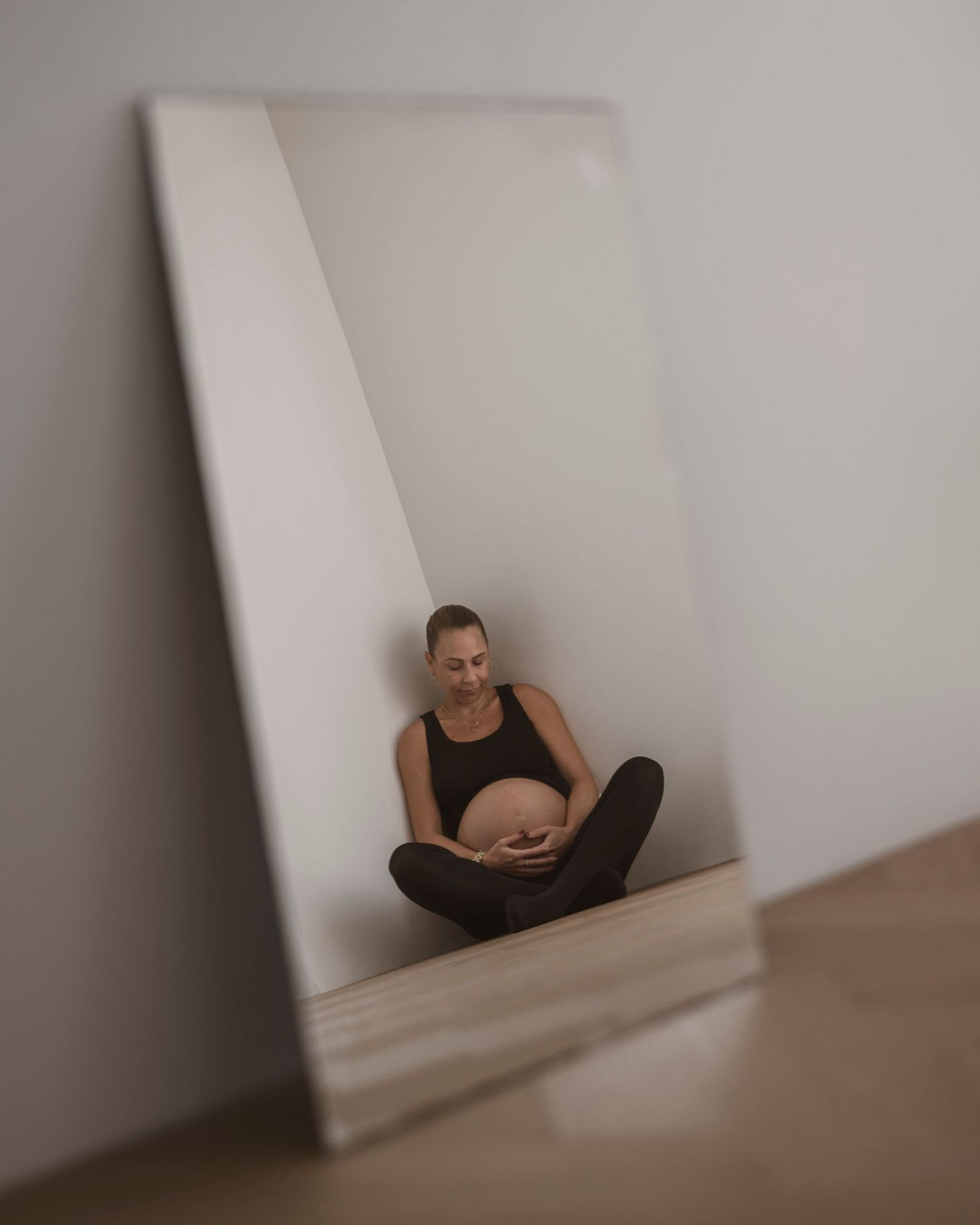 Pregnant woman sitting cross-legged cradling her baby bump, reflected in a large floor mirror against a white wall.