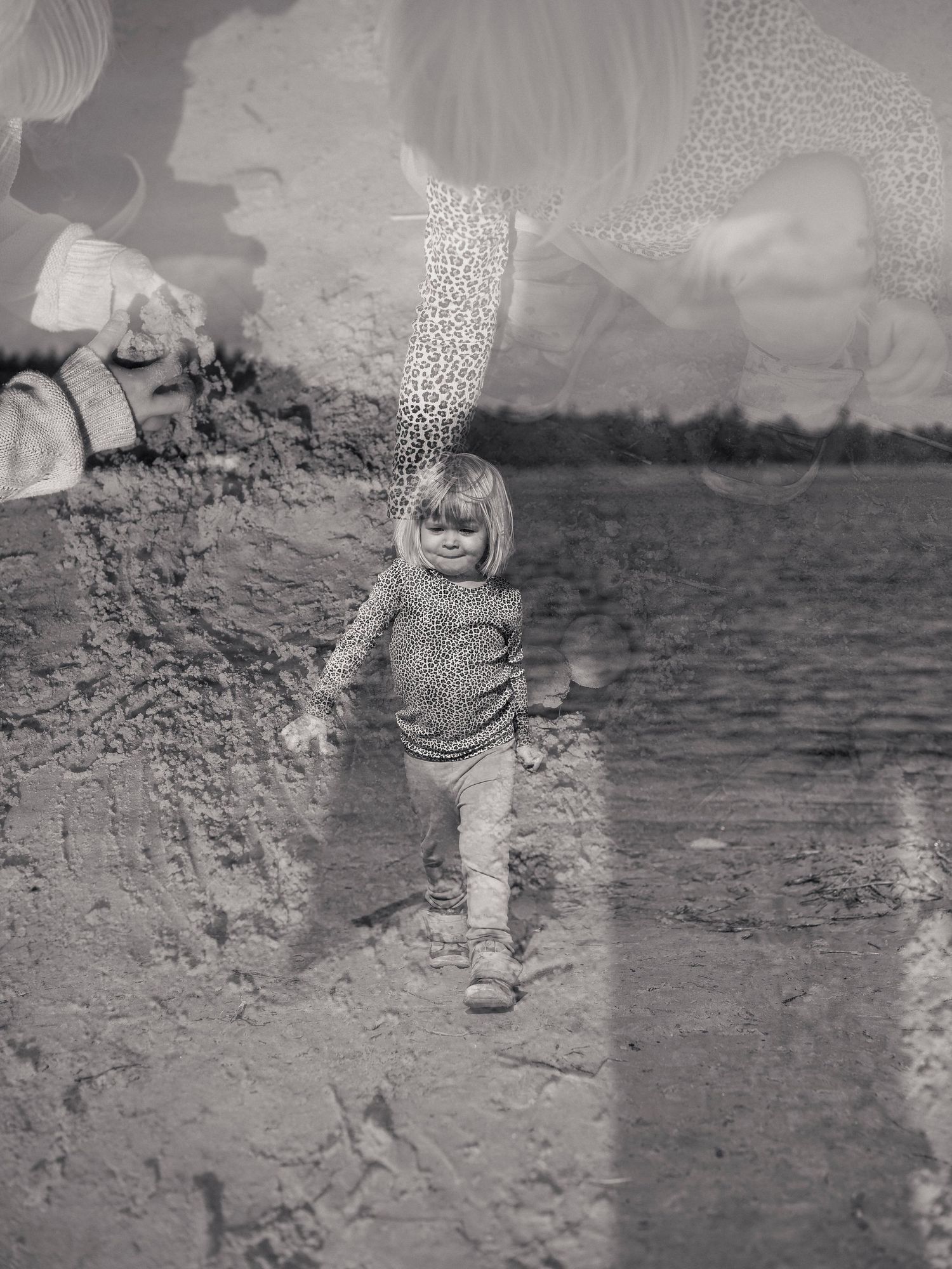 Black and white artistic double exposure of a toddler walking on a sandy beach near water.